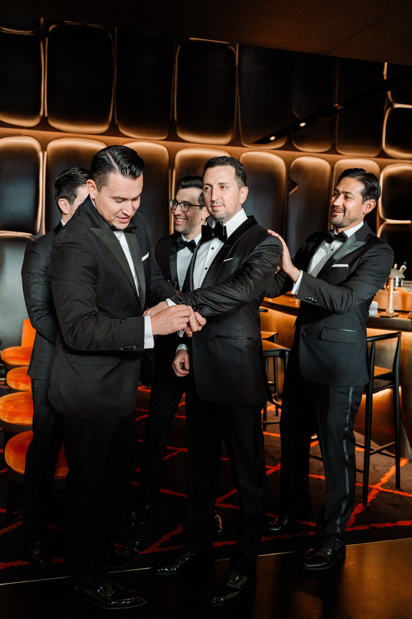 Groomsmen in tuxedos gather at modern bar with copper-toned lighting.