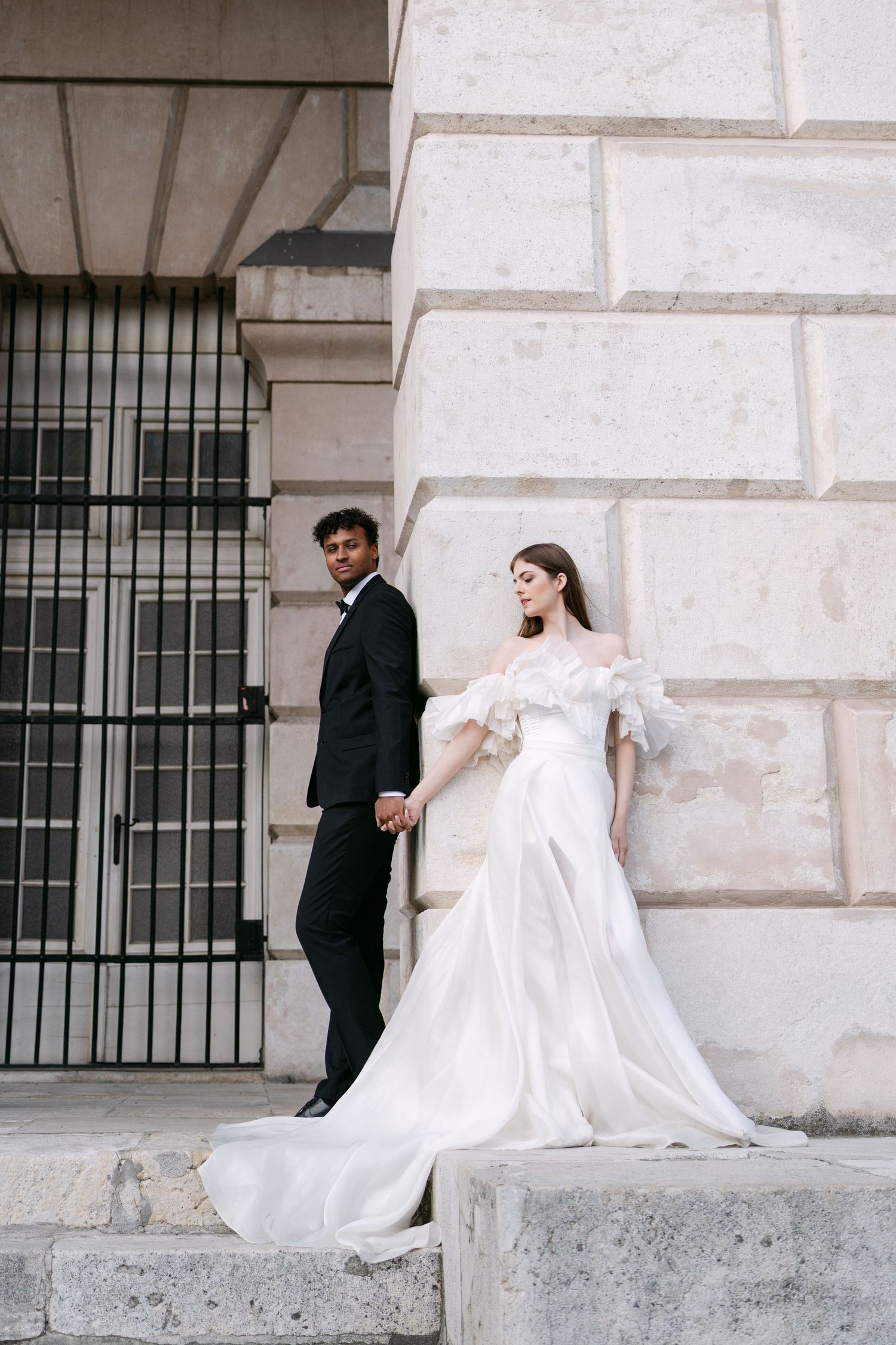 Couple in wedding attire poses dramatically against stone wall architecture.