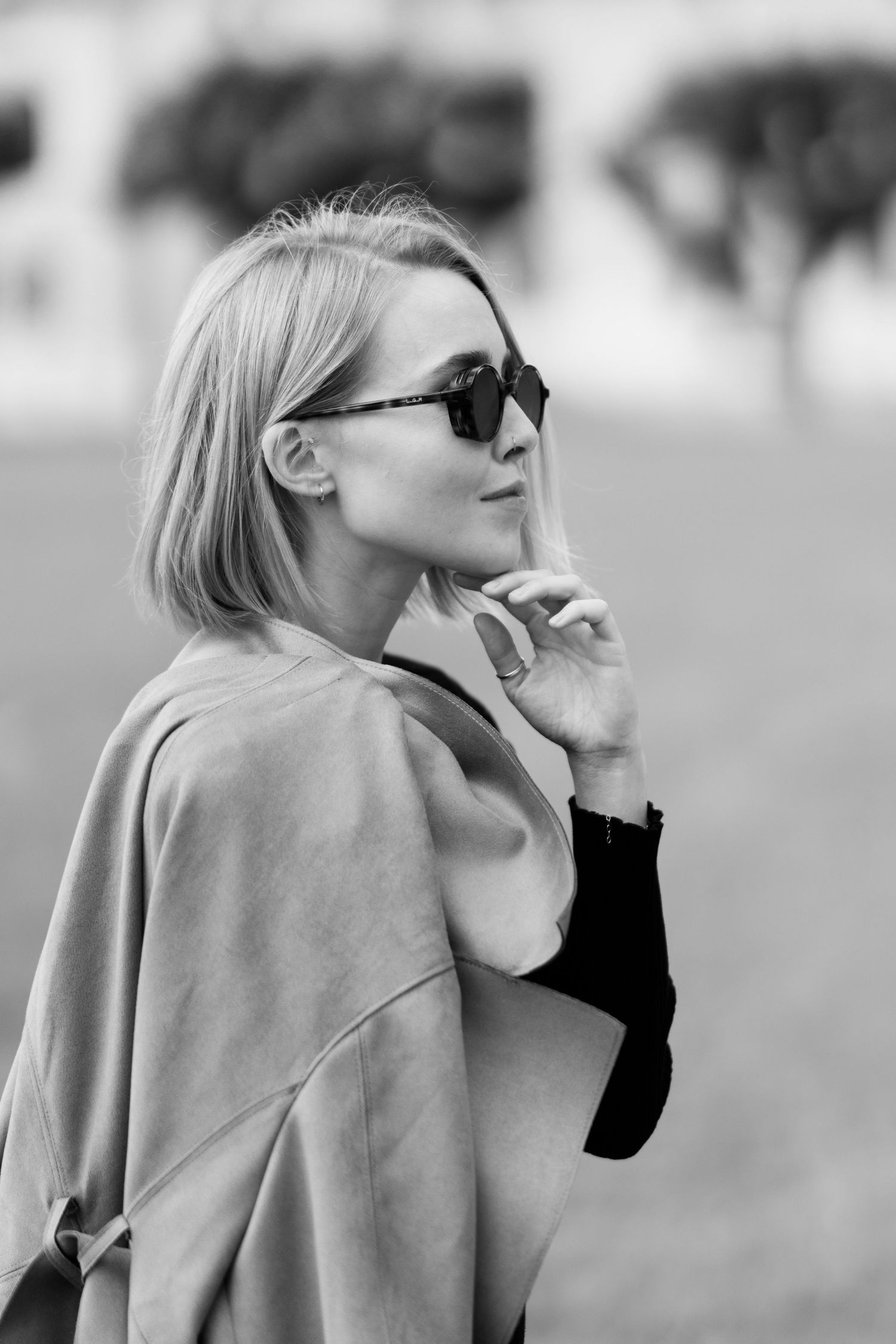 Black and white portrait of woman in sunglasses and coat with thoughtful expression.