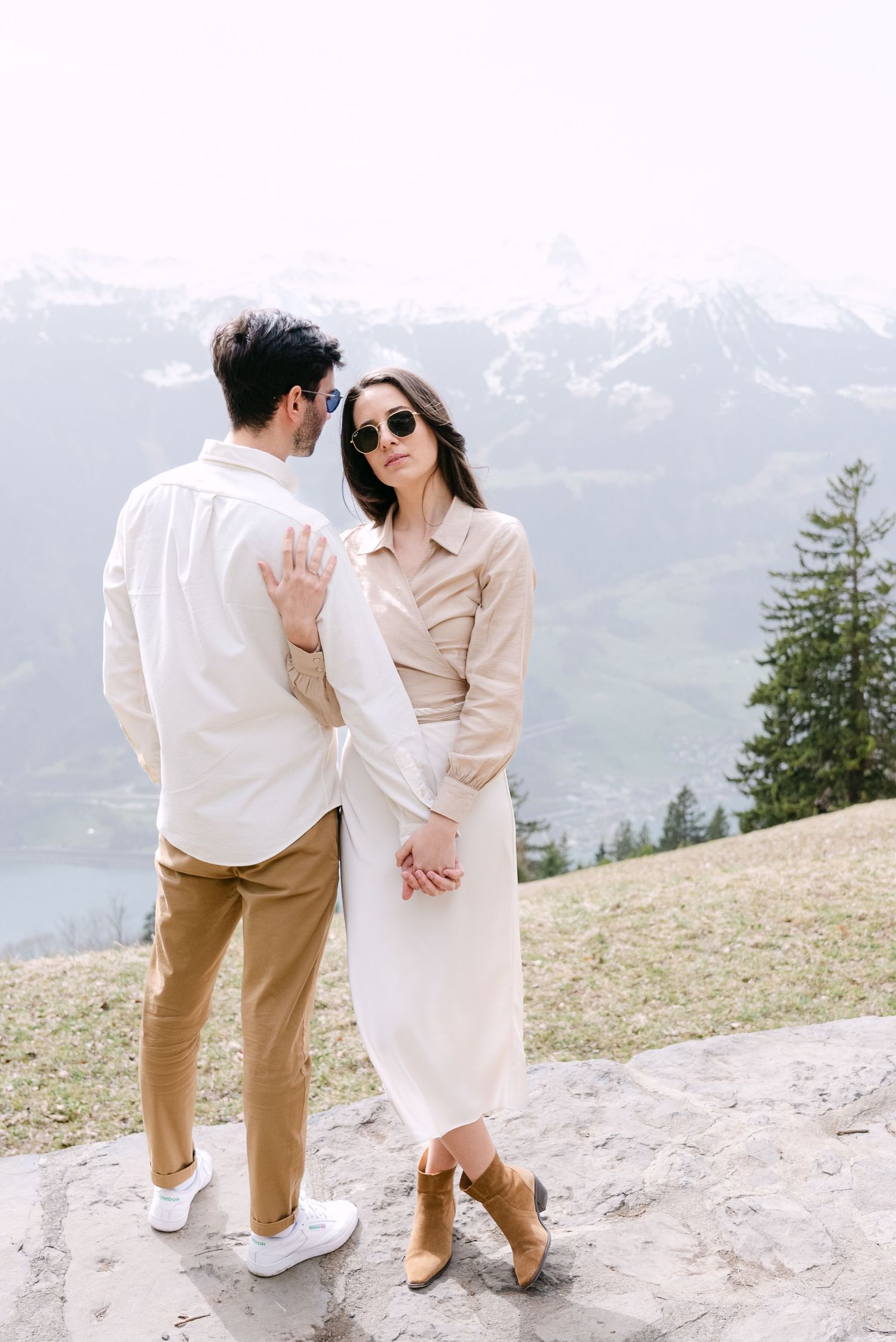 Couple in coordinated beige and white outfits pose romantically against snowy mountain backdrop.