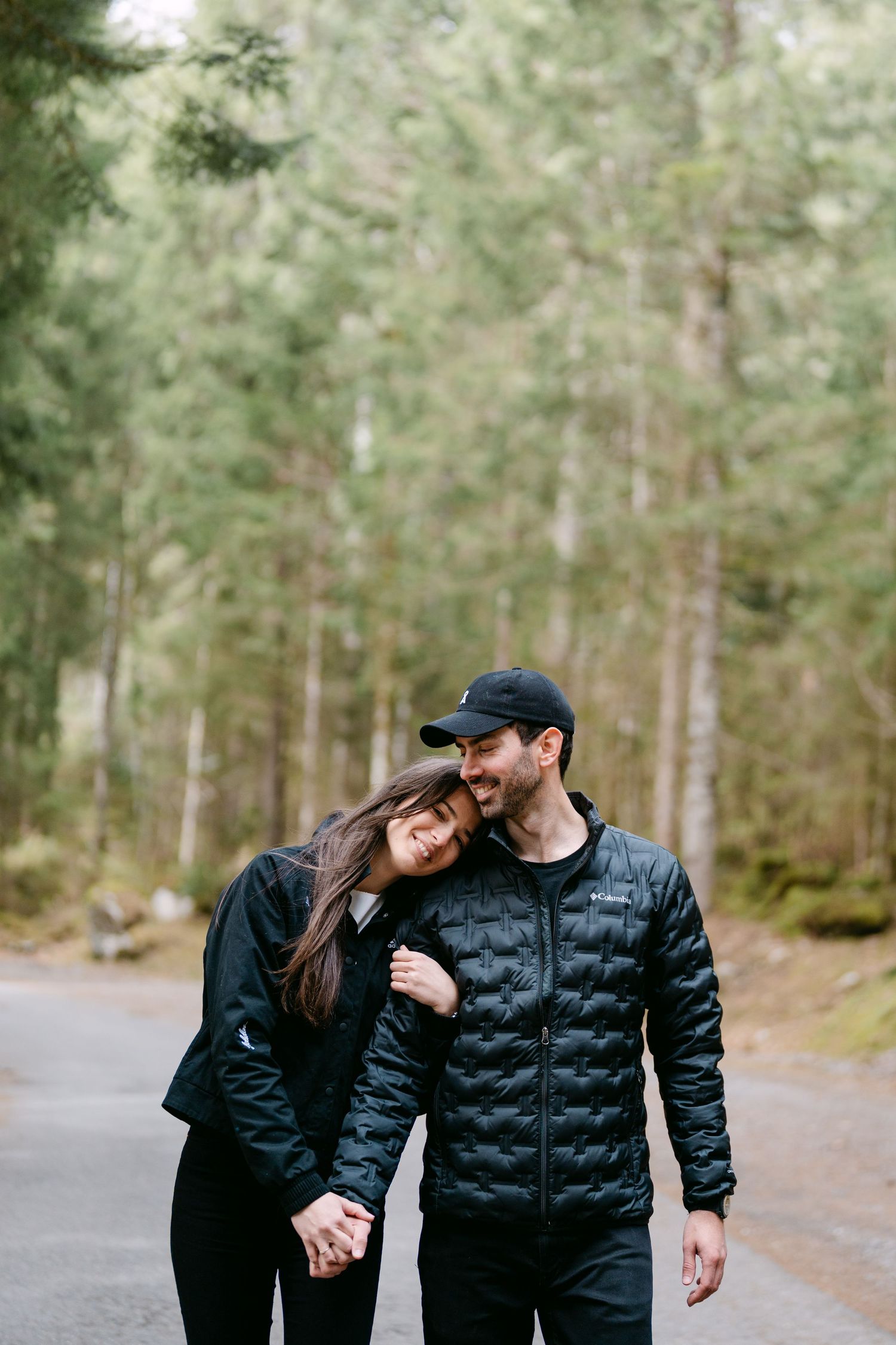Couple in matching black puffer jackets walking through a pine forest.