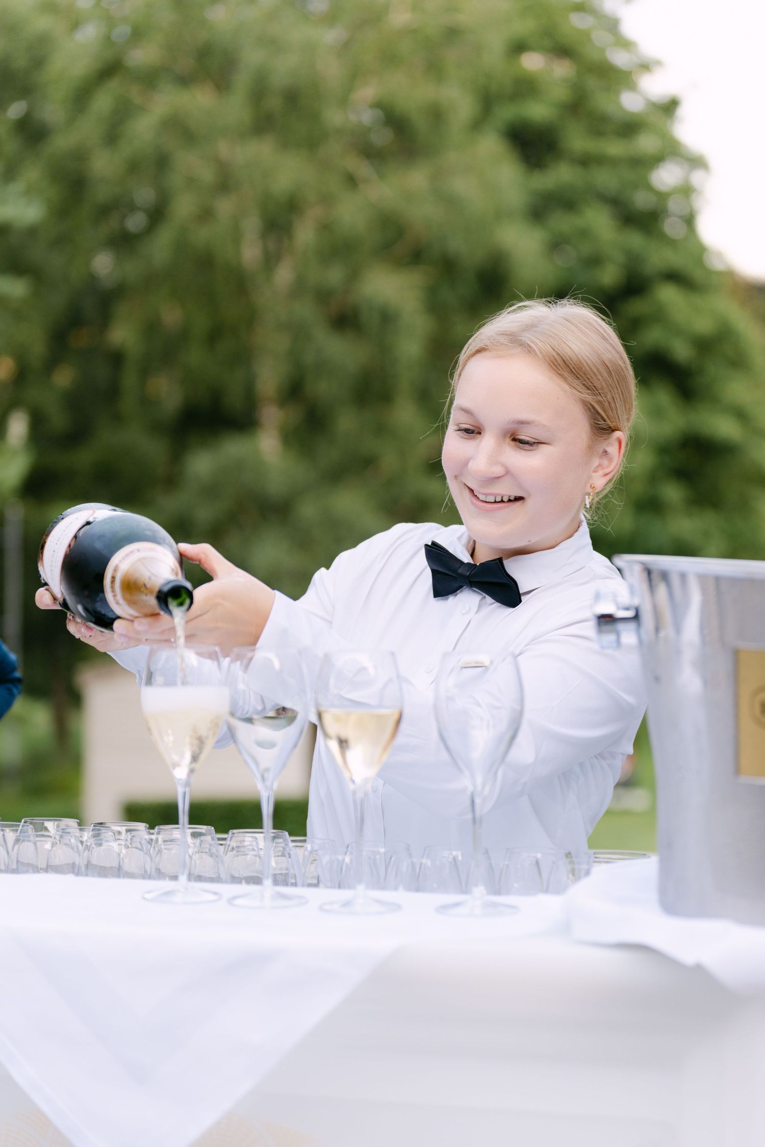 Server in white uniform carefully arranging glassware on a white-clothed table.