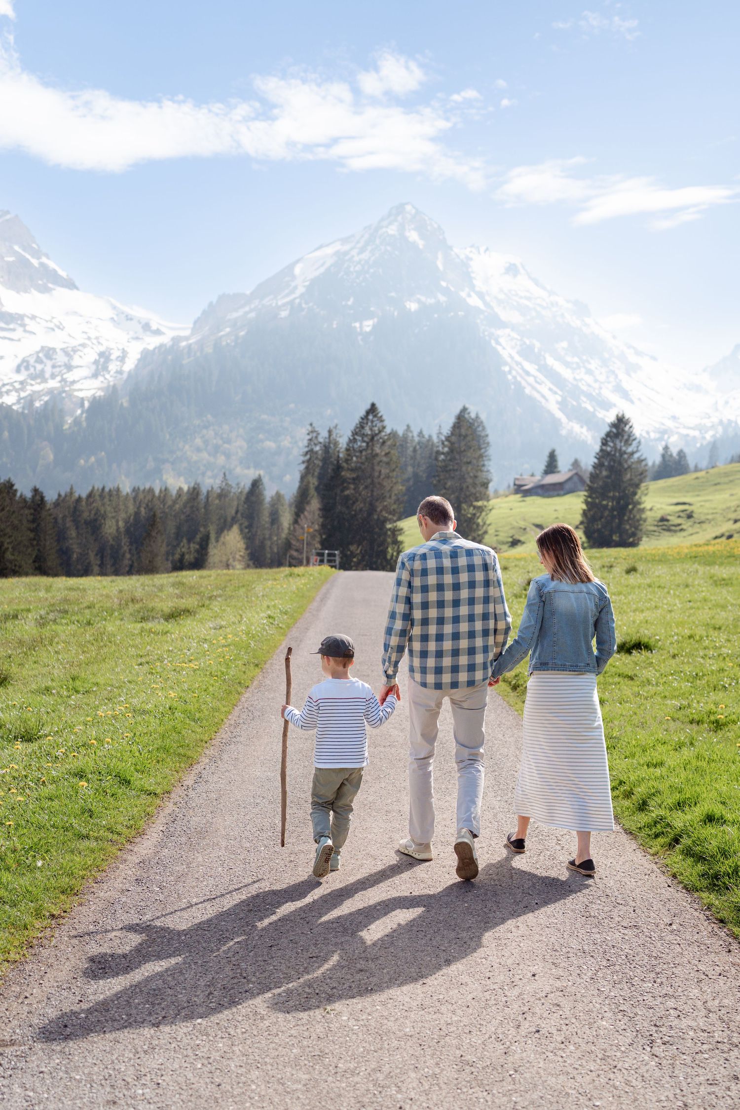 A family walks hand in hand down a mountain path with snow-capped peaks in view.