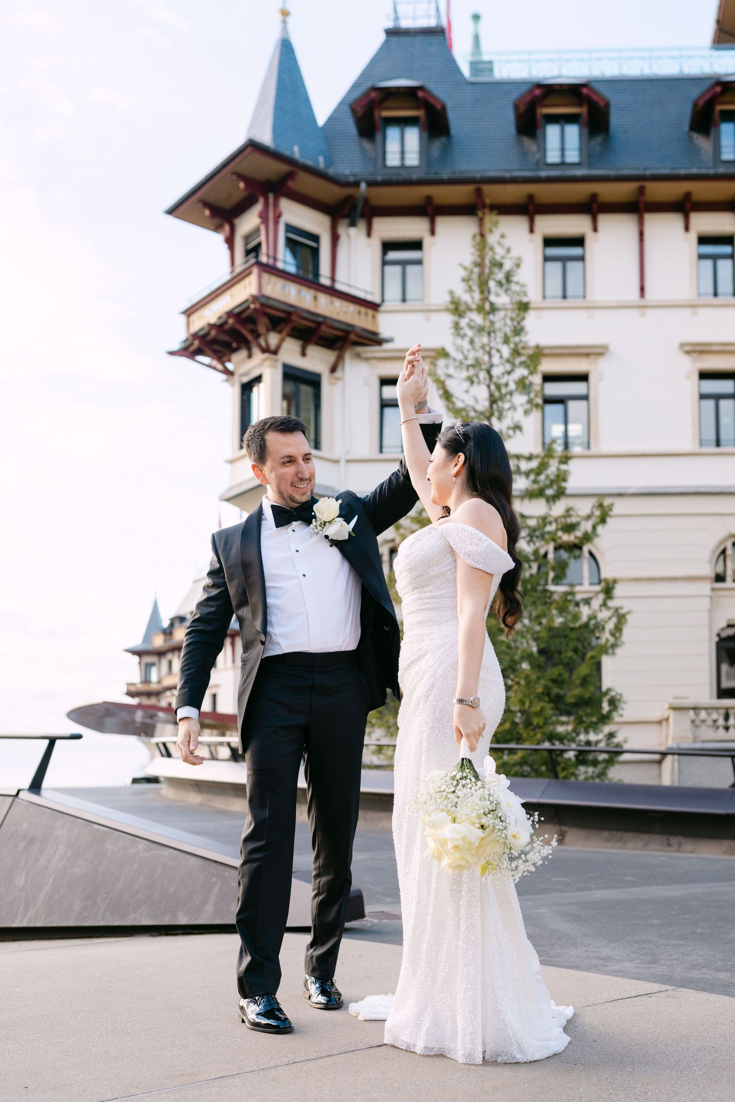 A couple in formal wedding attire poses outside a grand hotel building.