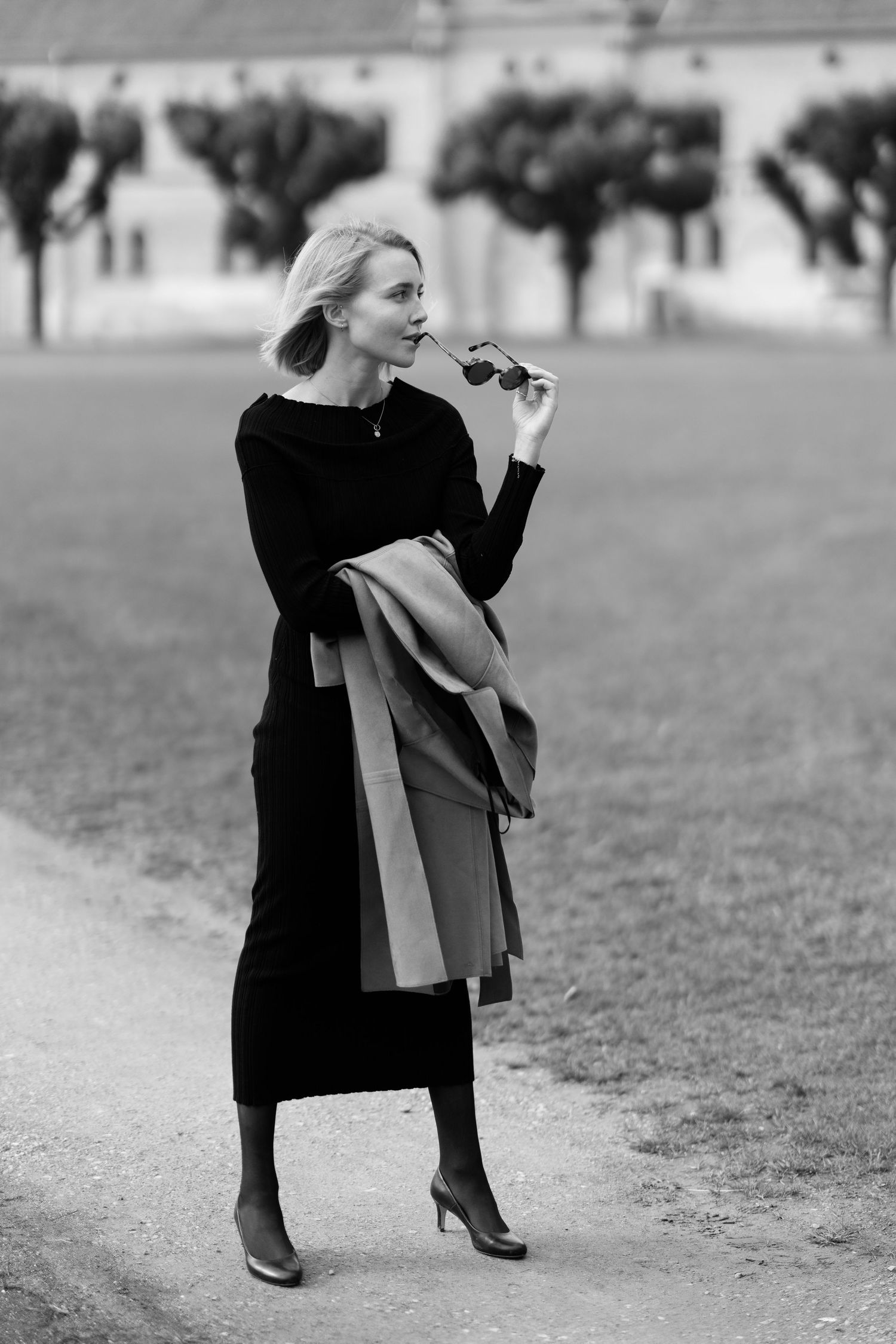 Black and white photo of woman in elegant black dress holding sunglasses outdoors.