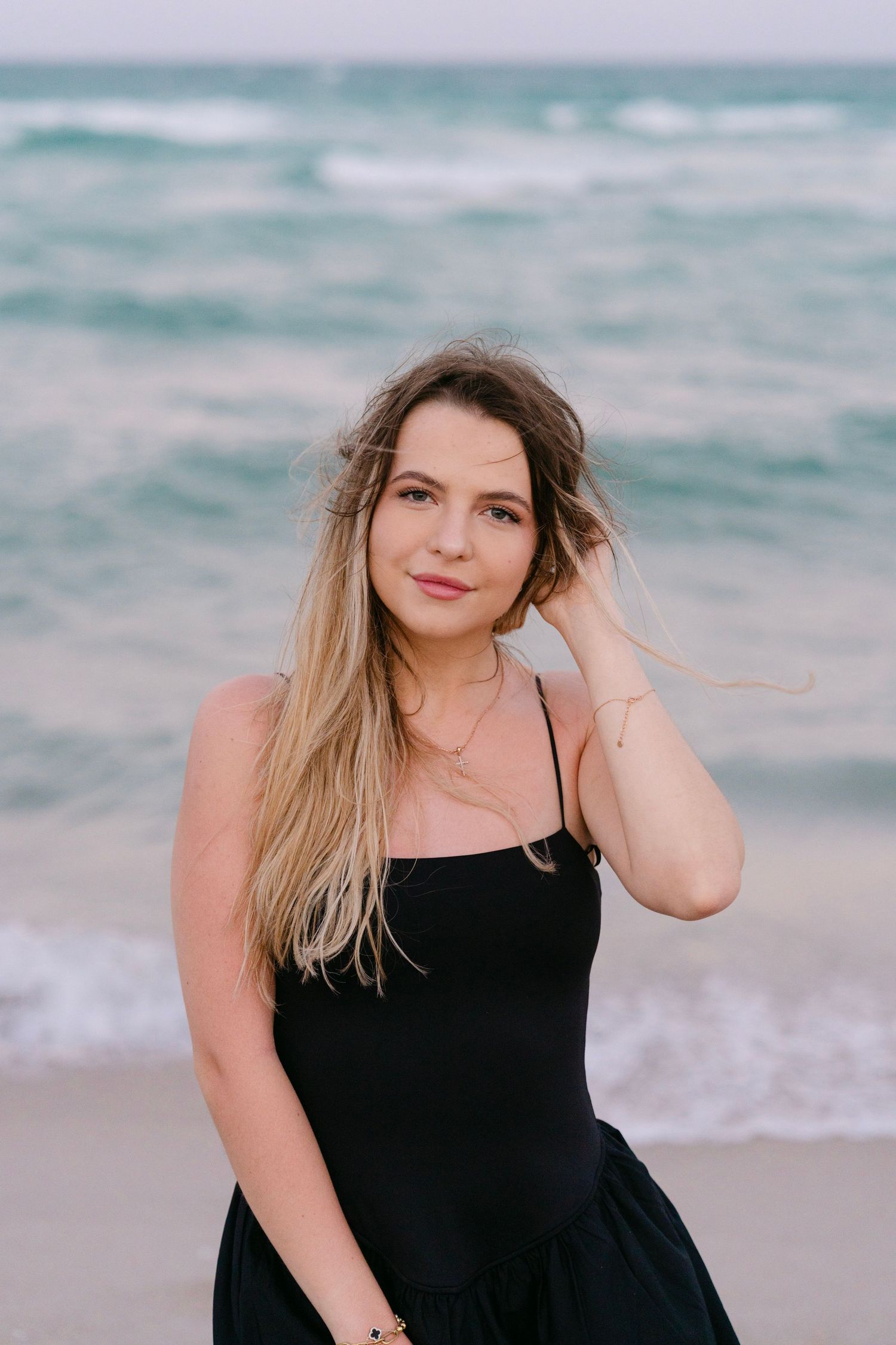 Woman in black dress poses on beach with windswept hair and ocean backdrop.