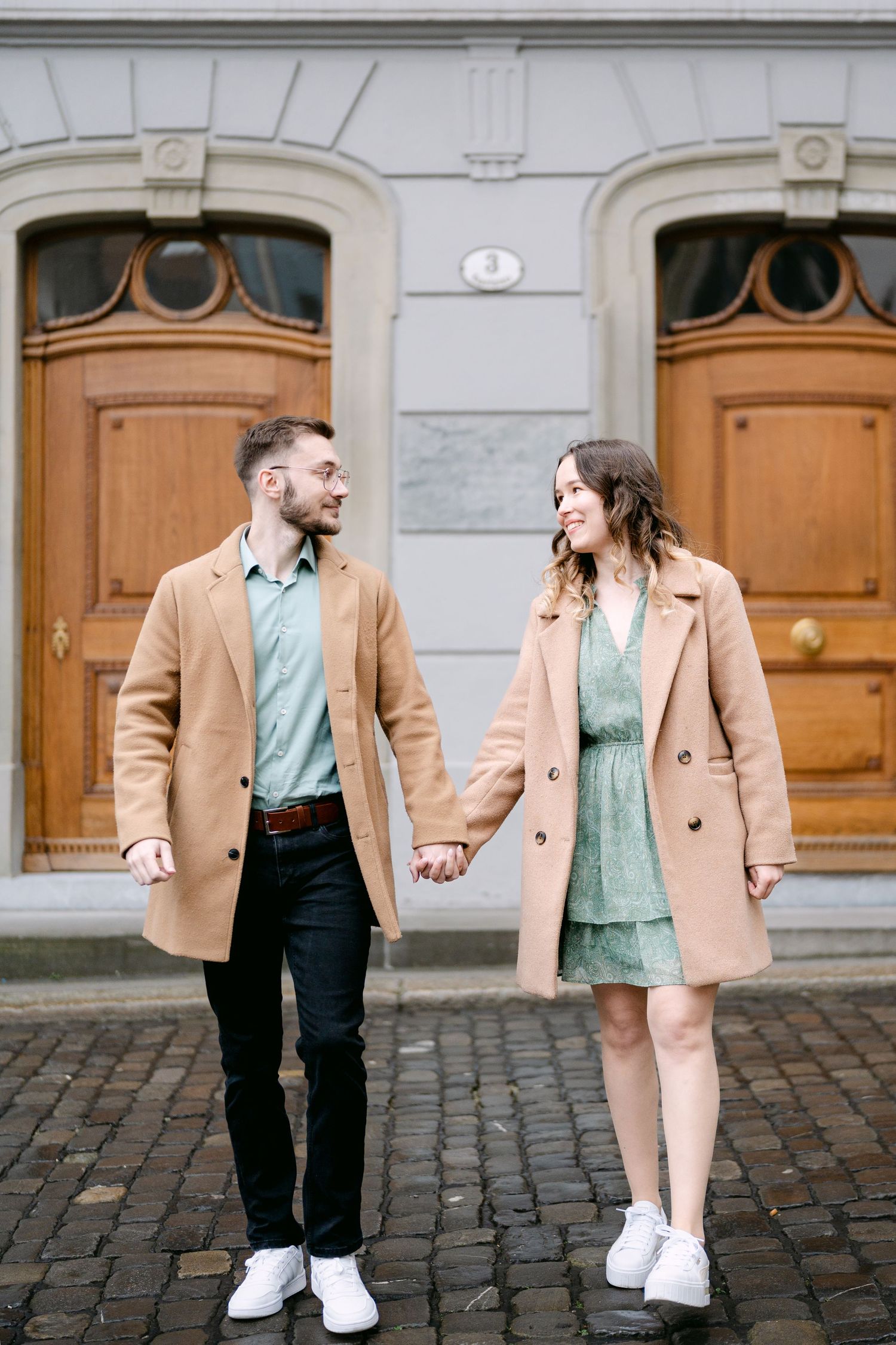 A stylish couple in camel coats and white sneakers pose on cobblestone streets.