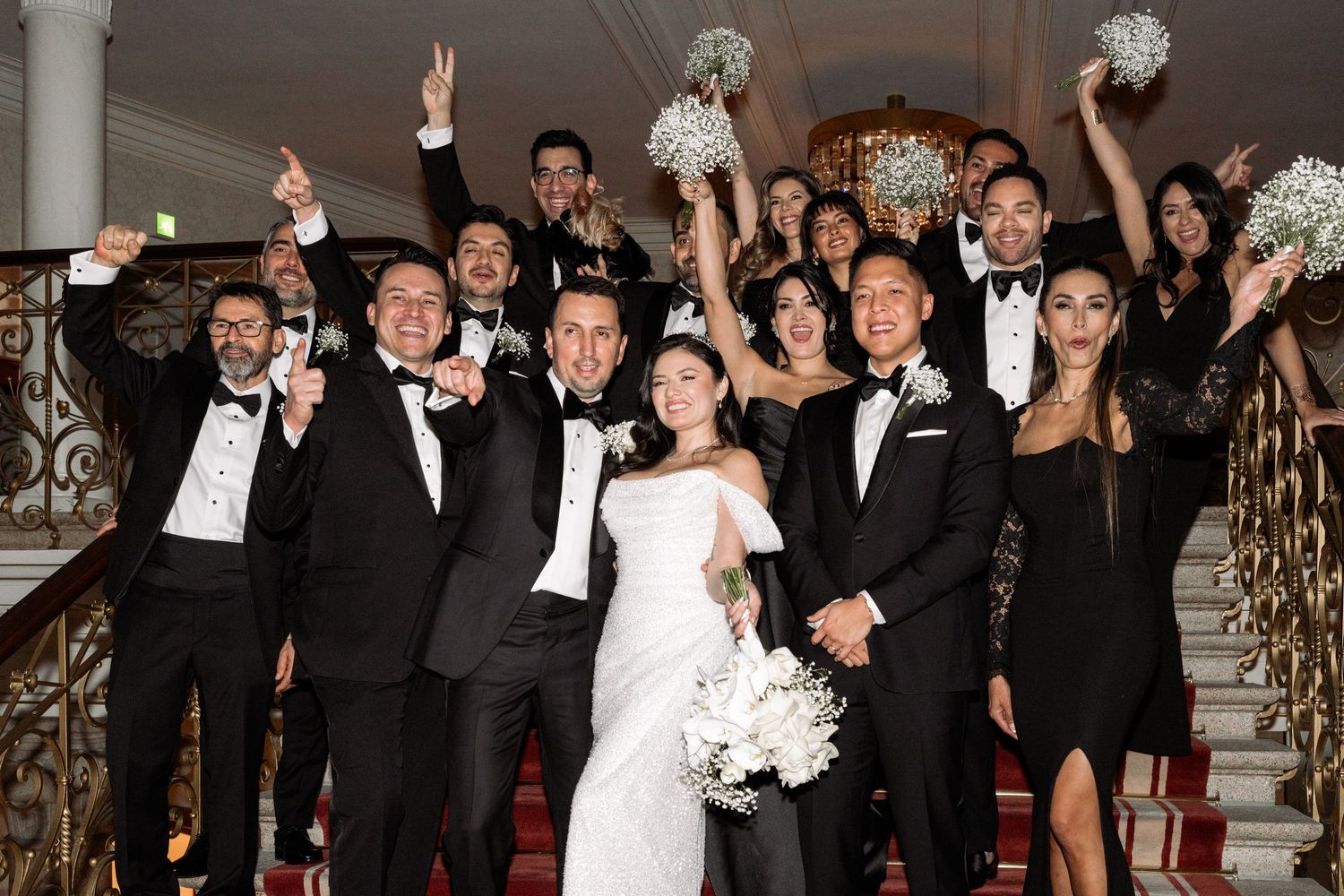 Wedding party celebrates joyfully on grand staircase with white bouquets and formal attire.