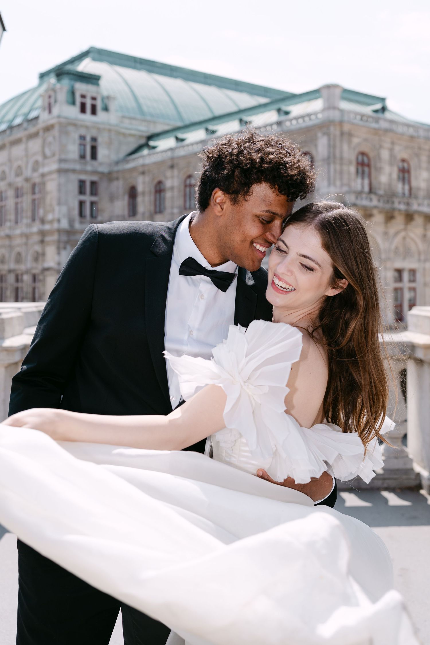 Joyful wedding couple embraces on balcony of historic Vienna Opera House.