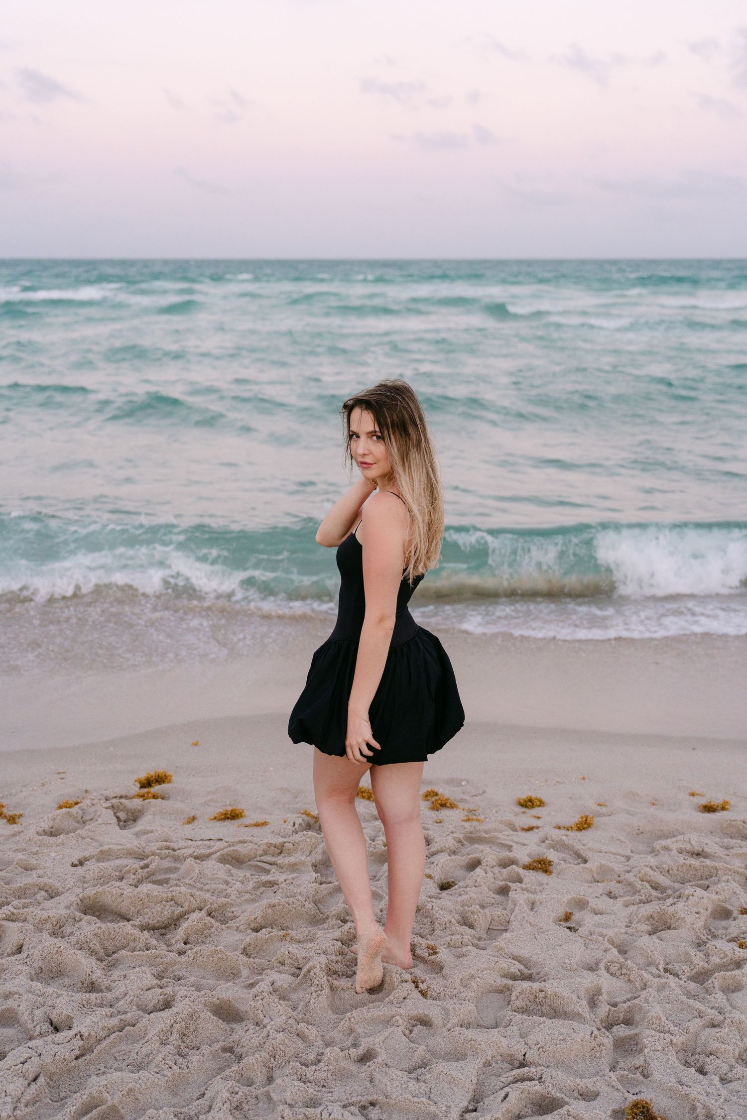 A woman in a black dress poses playfully on a sandy beach with turquoise ocean waves in the background.