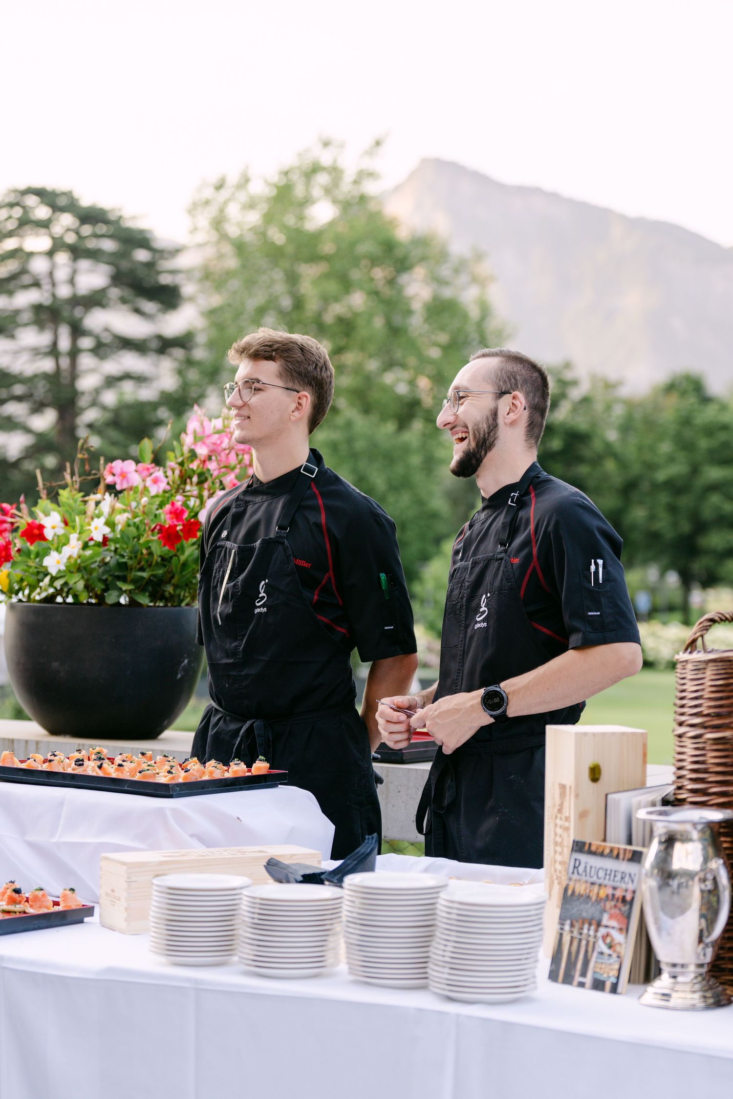 Catering staff in black uniforms prepare food at an outdoor buffet table with white linens and stacked plates.