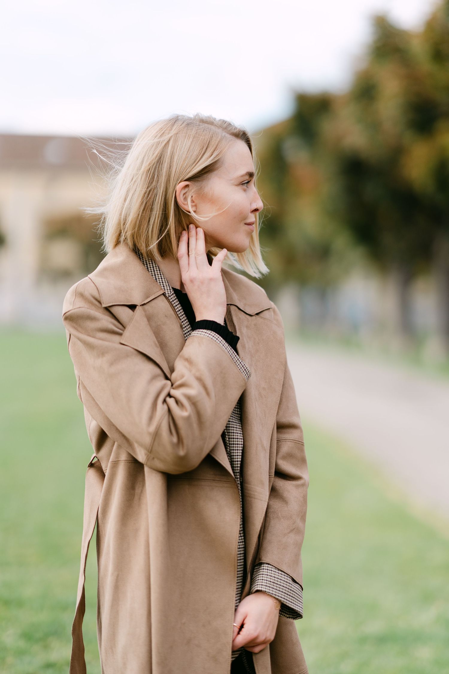 Fashionable woman in beige coat poses outdoors on path with autumn foliage in background.