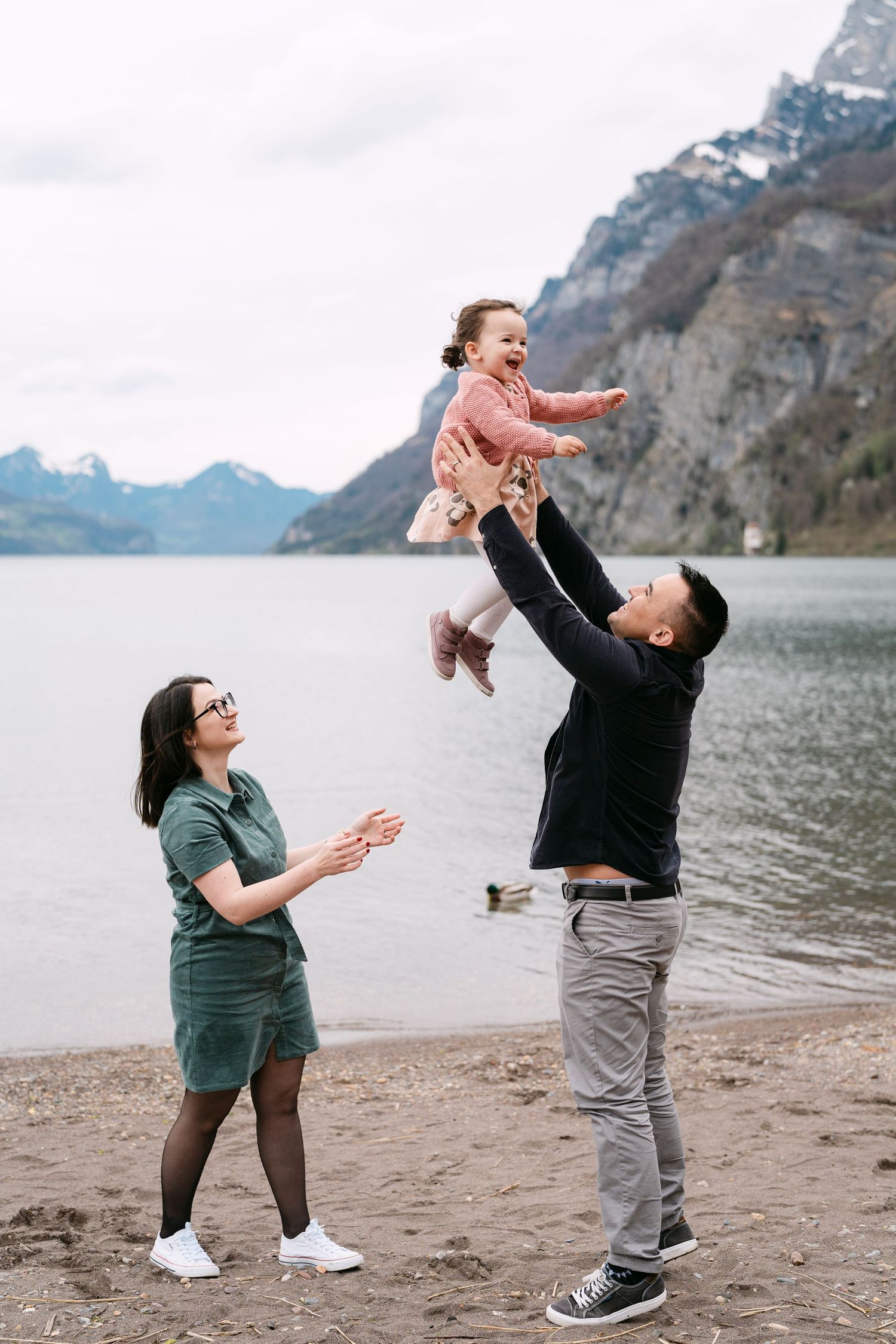 A playful family moment captured on a beach with snow-capped mountains in the background.