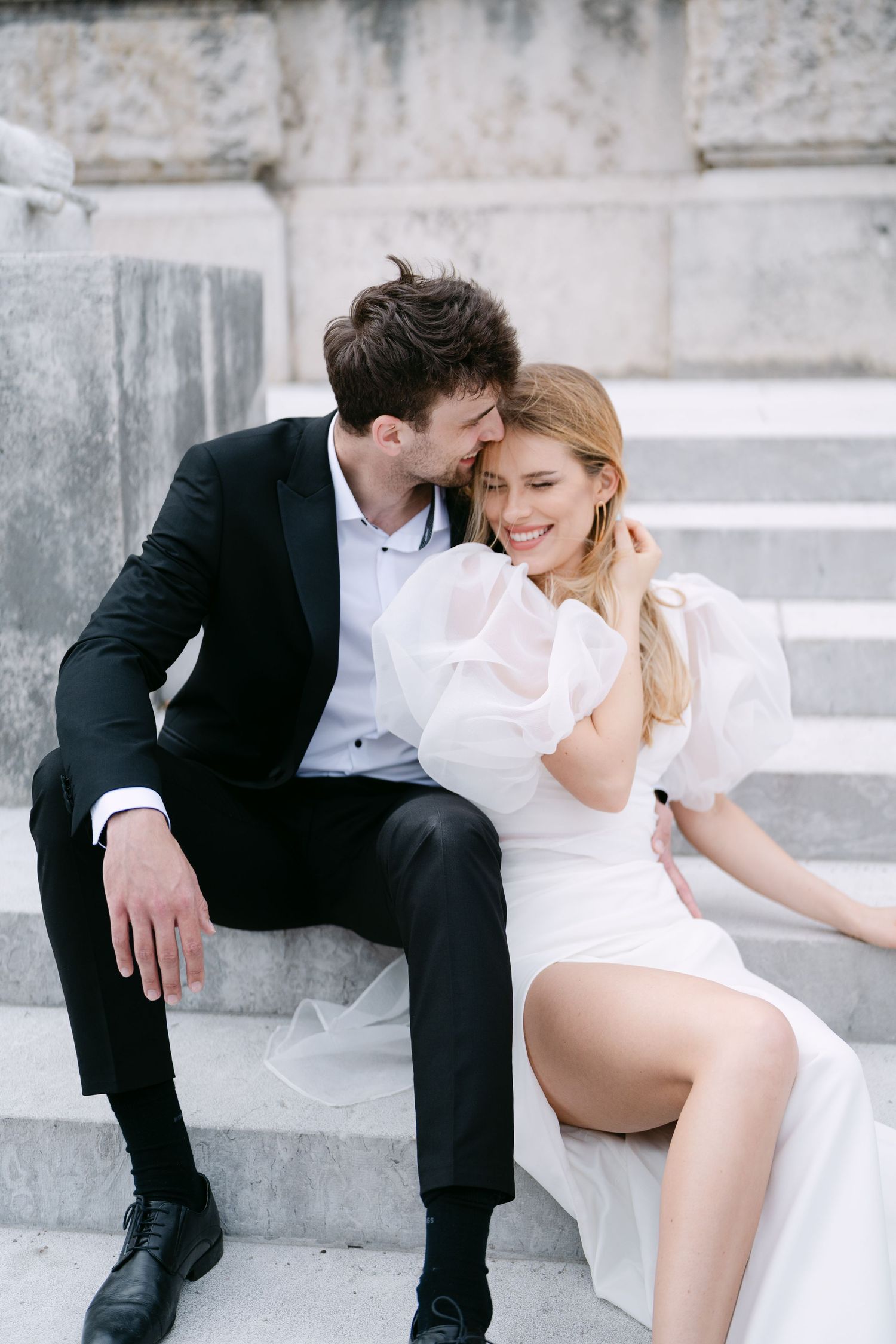 Romantic couple sitting on marble steps in elegant wedding attire.