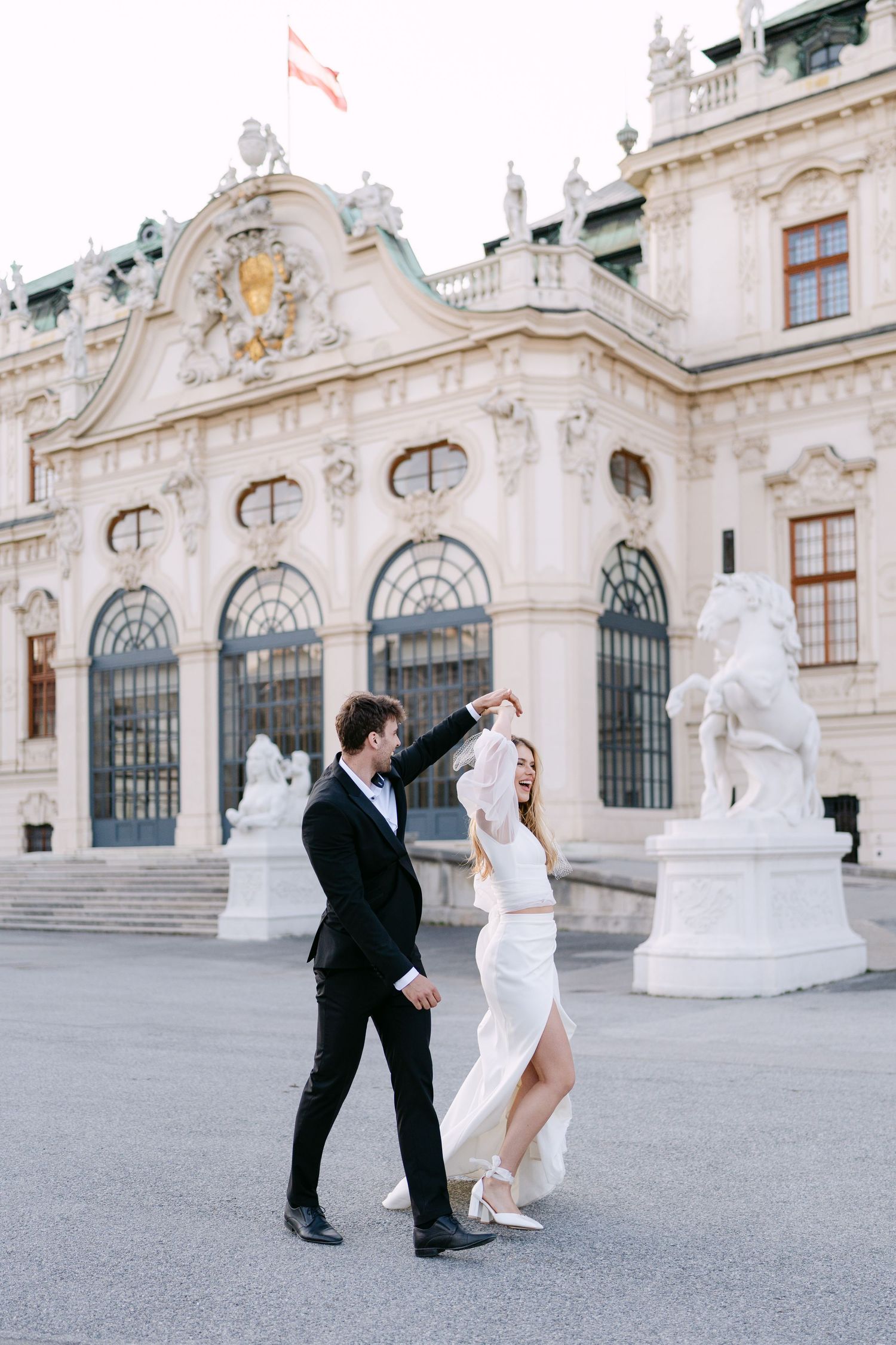 Wedding couple dances playfully in front of ornate Baroque palace facade.