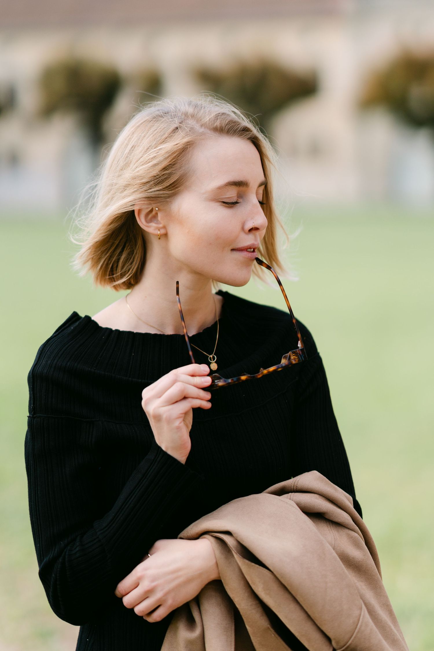 Woman in black sweater holding tortoise shell sunglasses against blurred architectural background.
