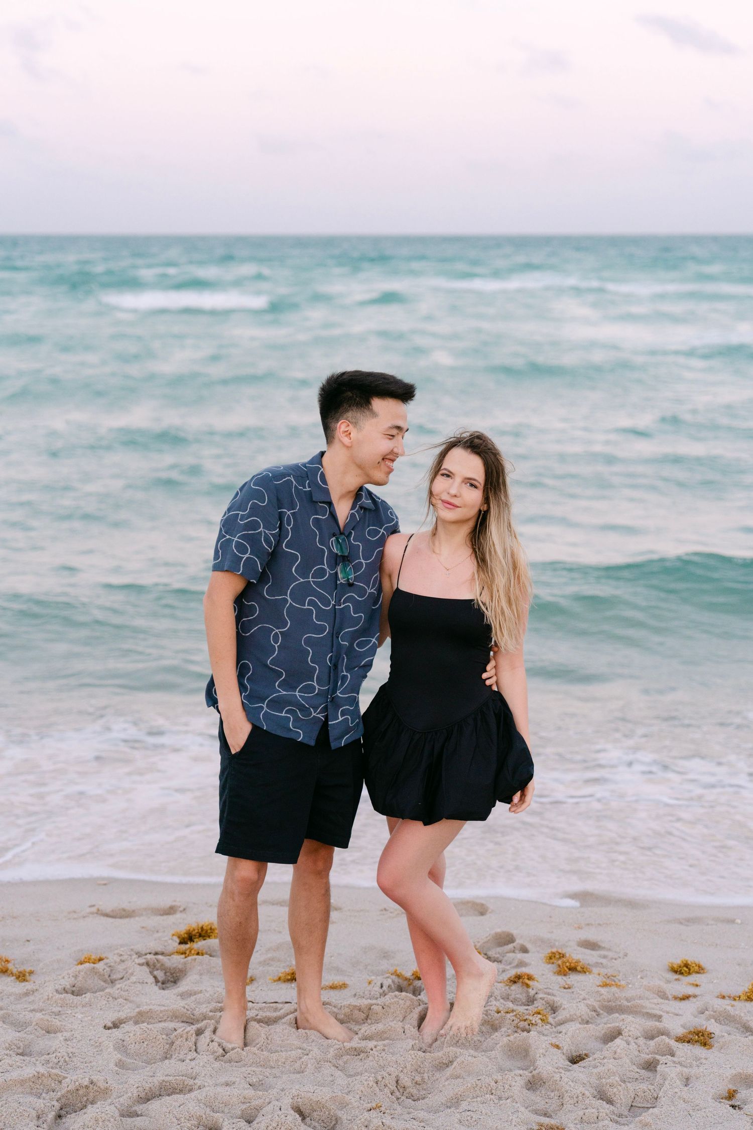 Two people laugh together while posing casually on a sandy shoreline.
