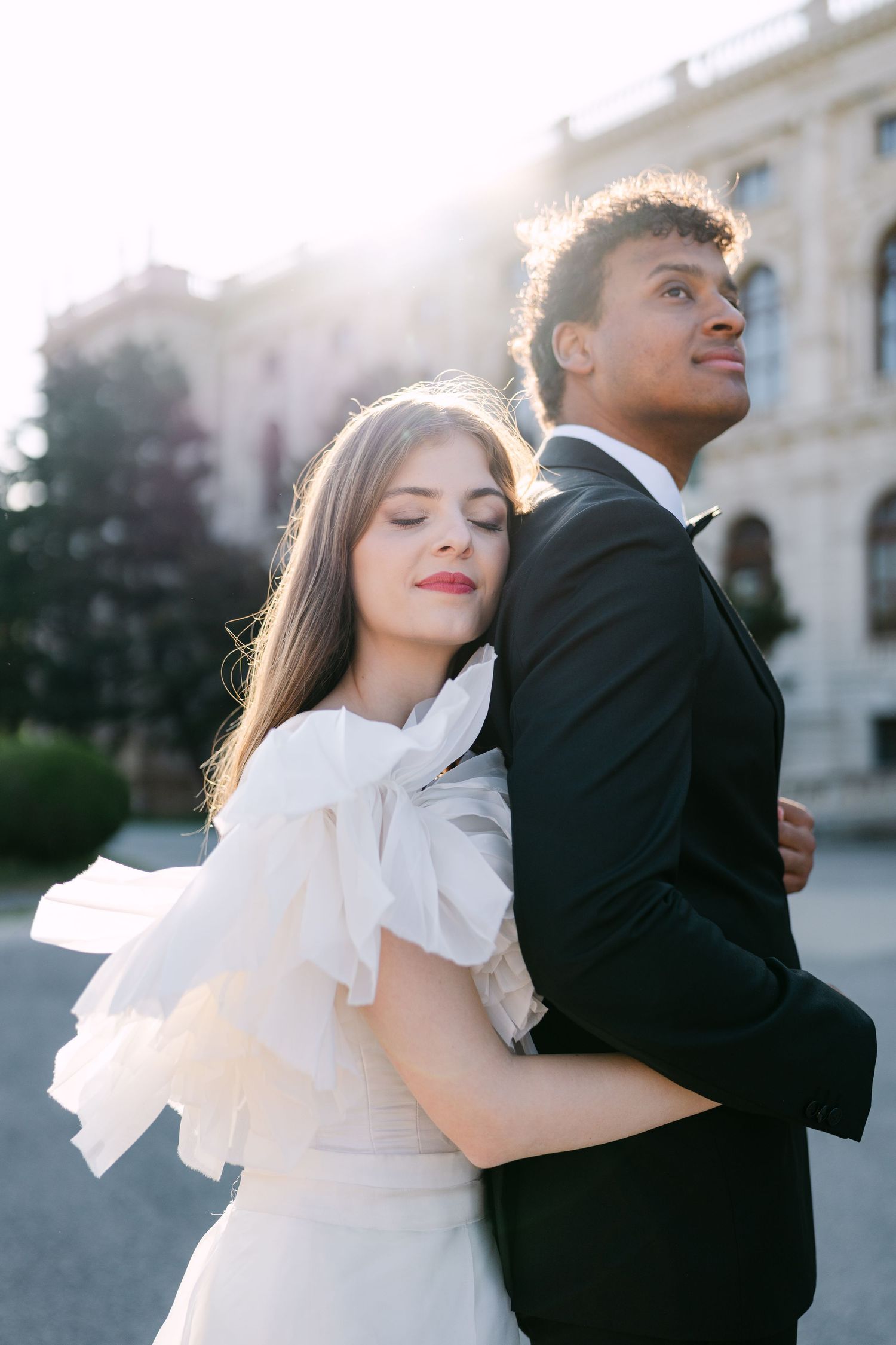 Romantic couple in wedding attire share intimate moment against classic architecture backdrop.