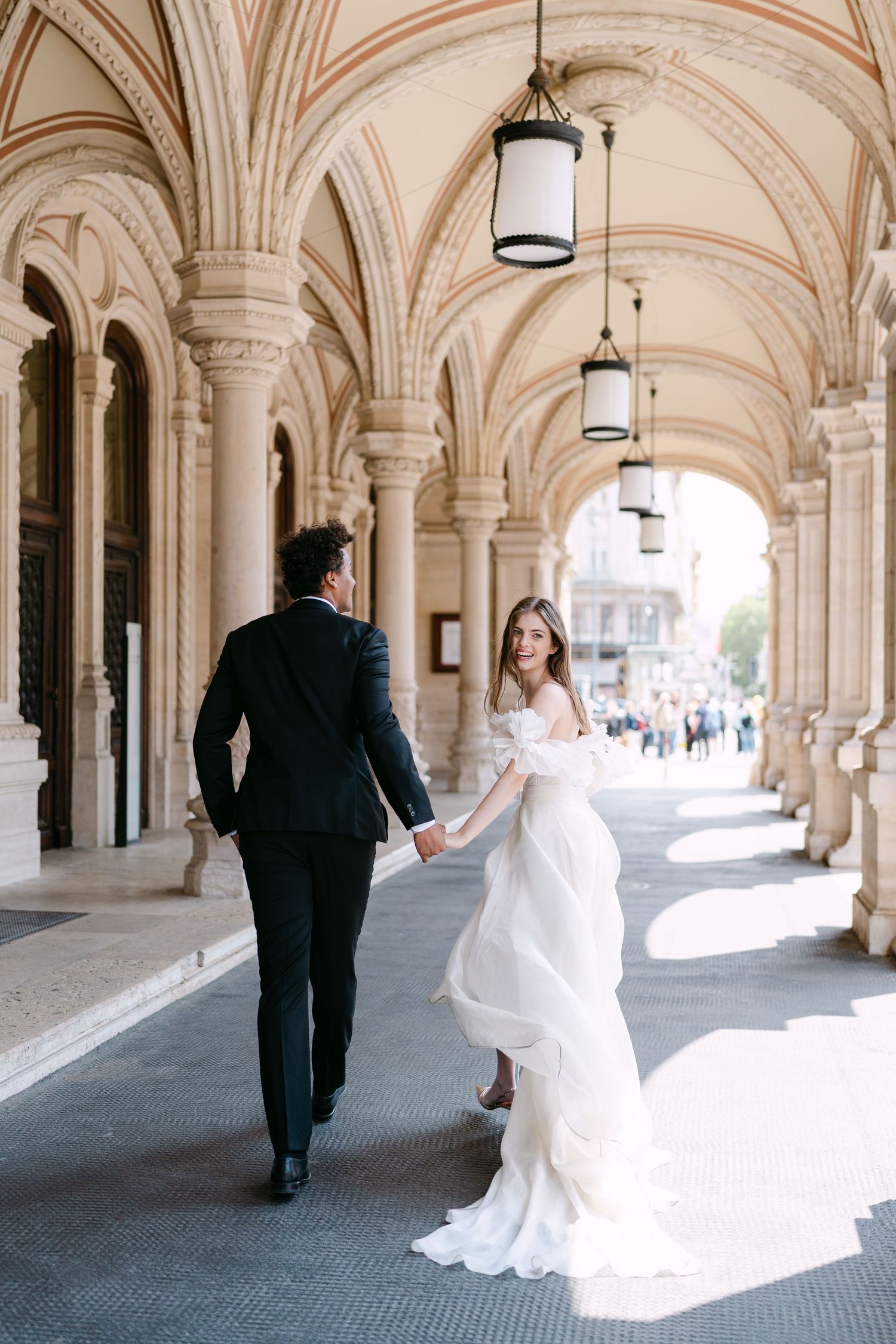 Elegant wedding couple walks through ornate stone archways with flowing white dress trailing behind.
