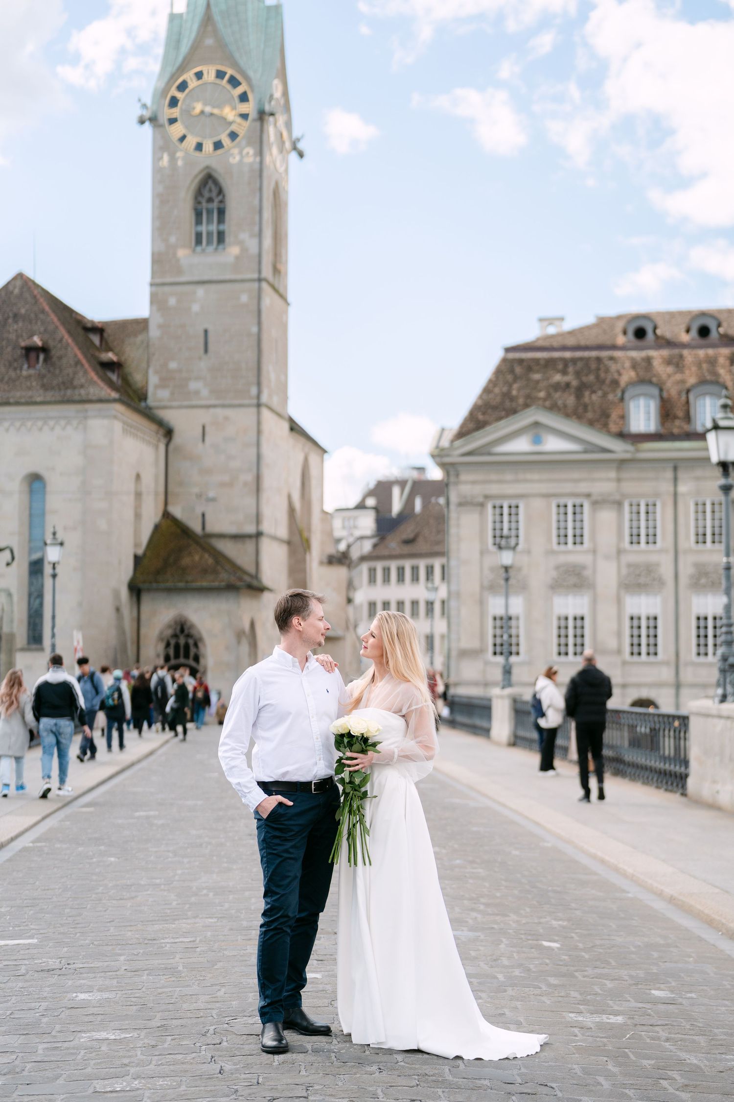 Wedding couple poses on historic European plaza with church tower in background.