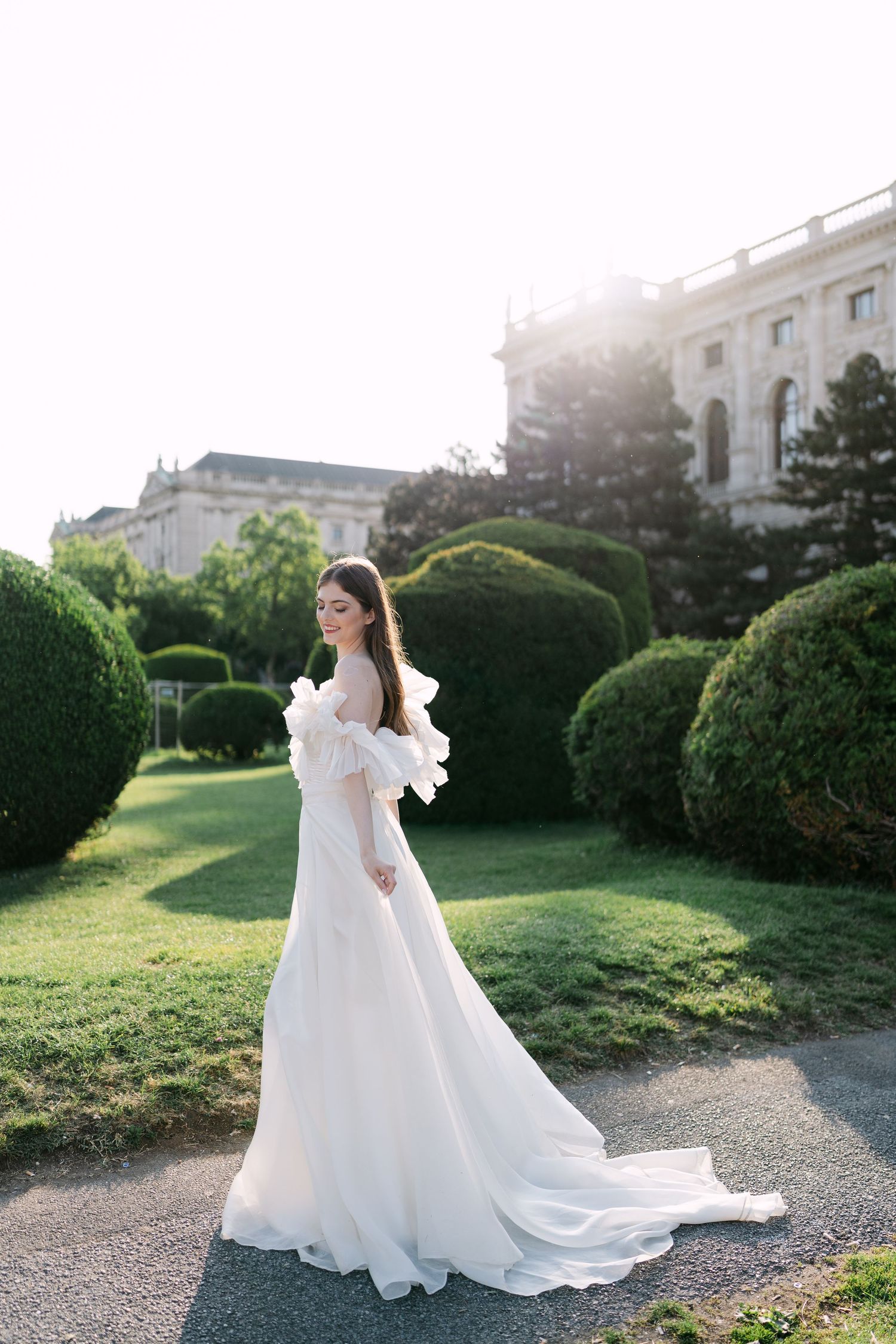 Bride in flowing white gown poses in garden setting with classical architecture.