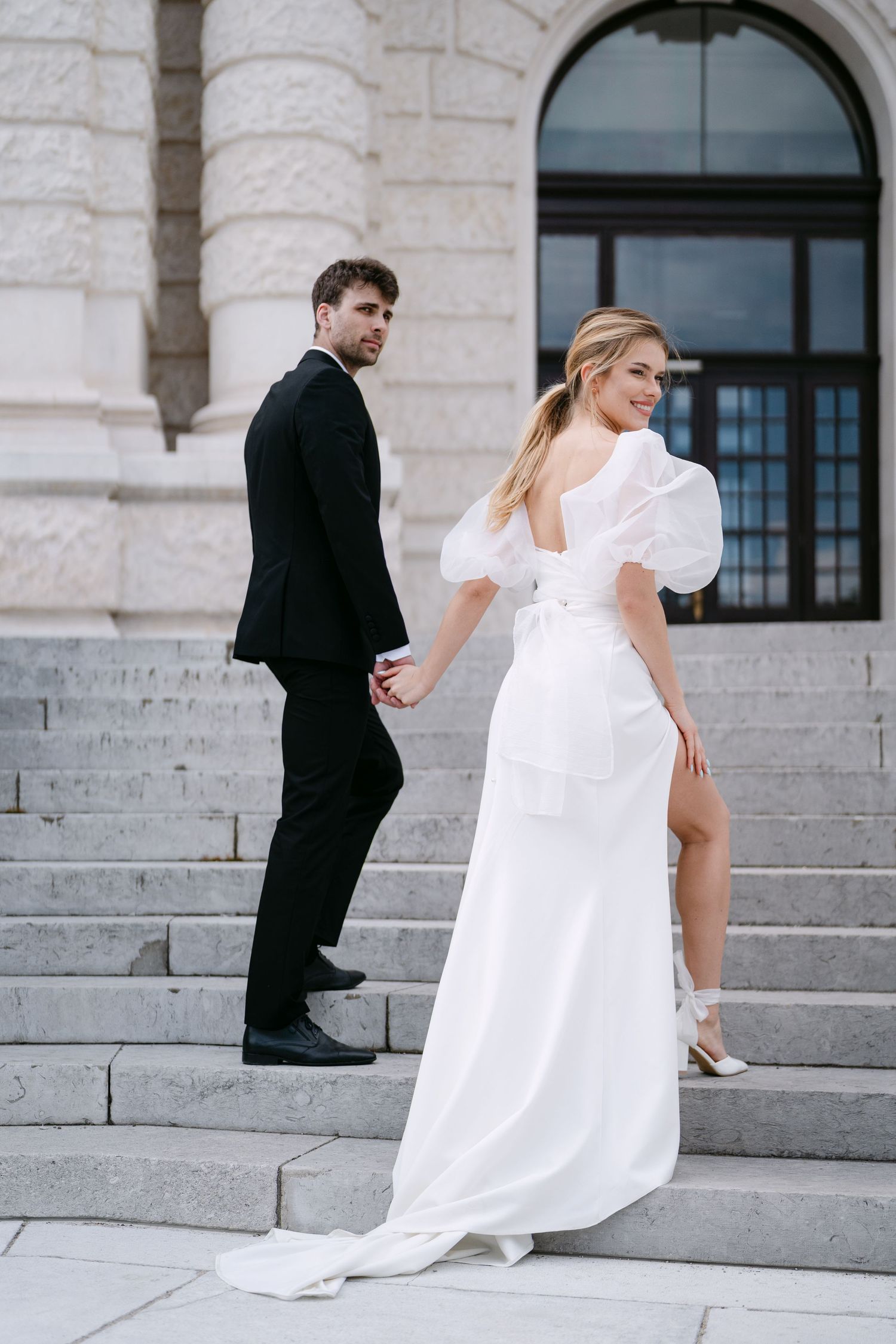 Wedding couple ascending ornate stone steps with flowing white dress and classic tuxedo.