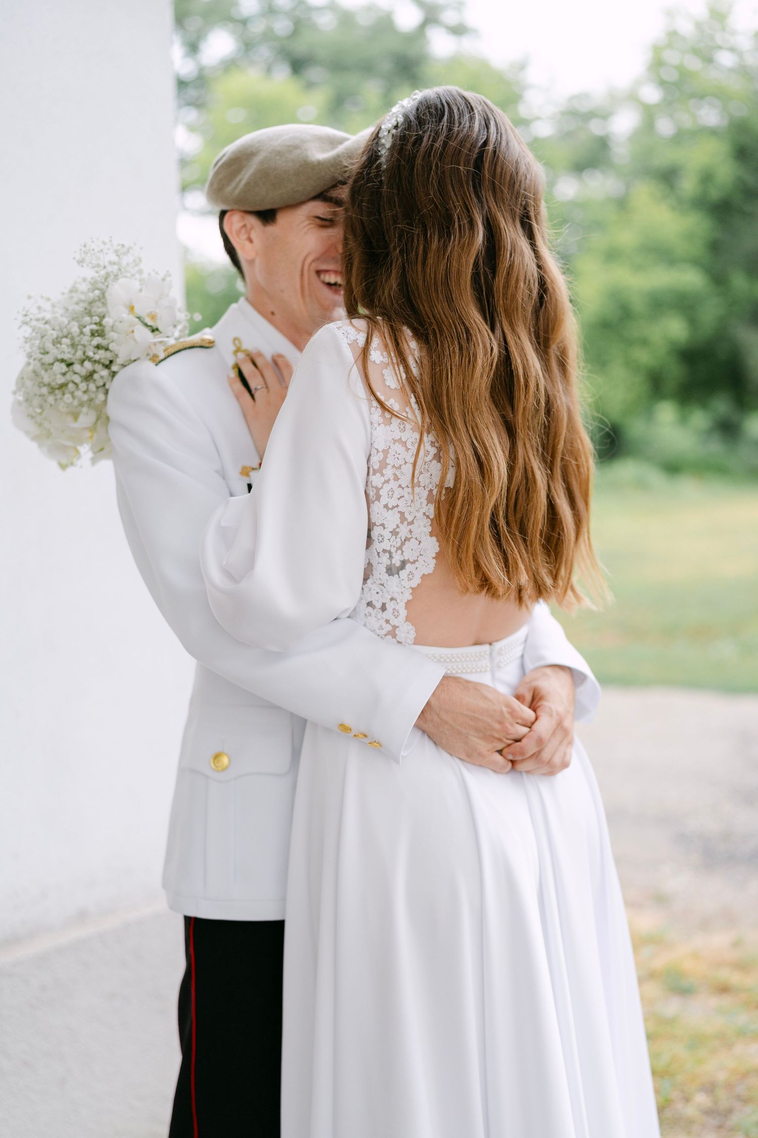 Military couple in white formal attire embrace outdoors with baby's breath flowers.