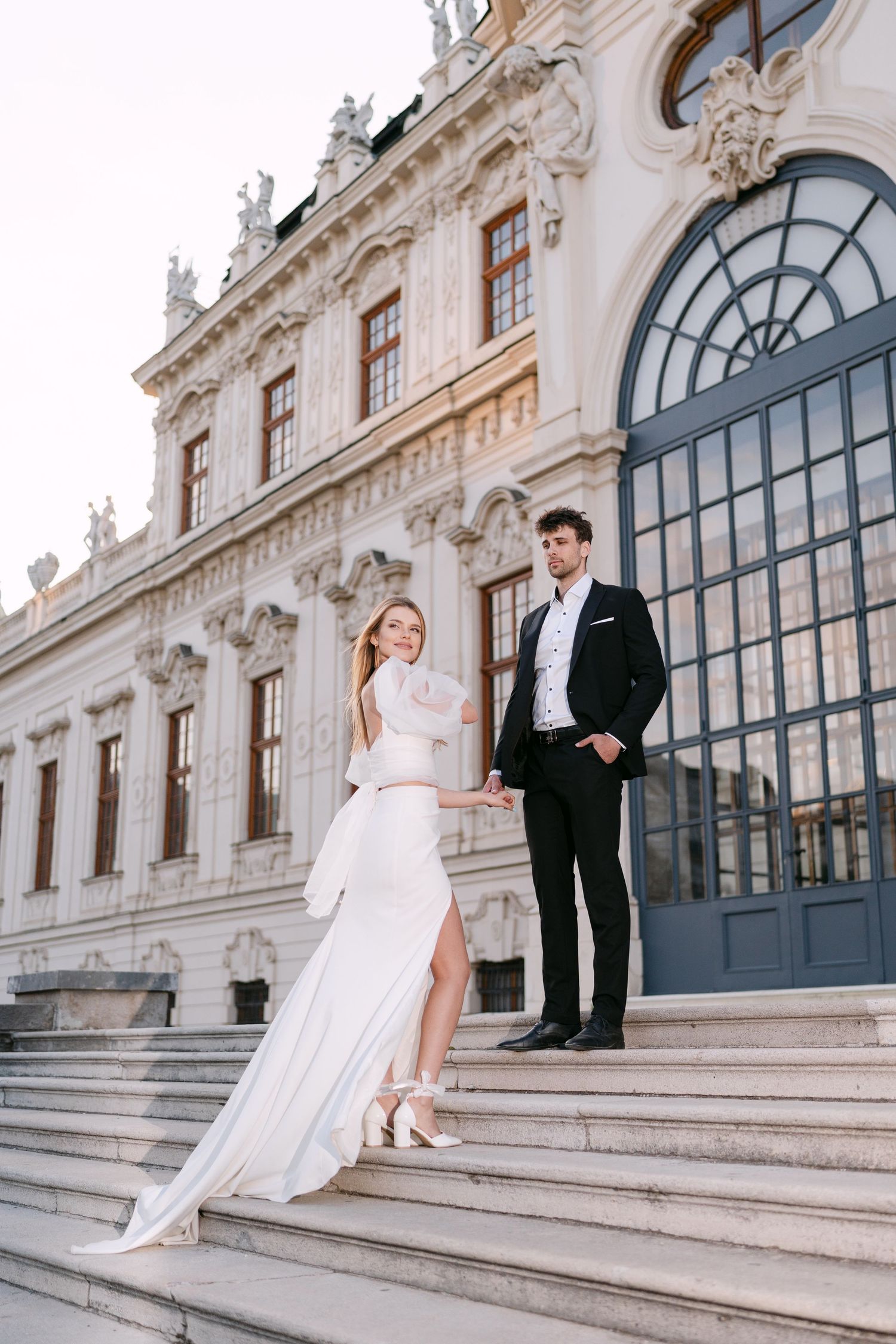 Romantic wedding photos captured on the steps of a historic baroque building with arched windows.