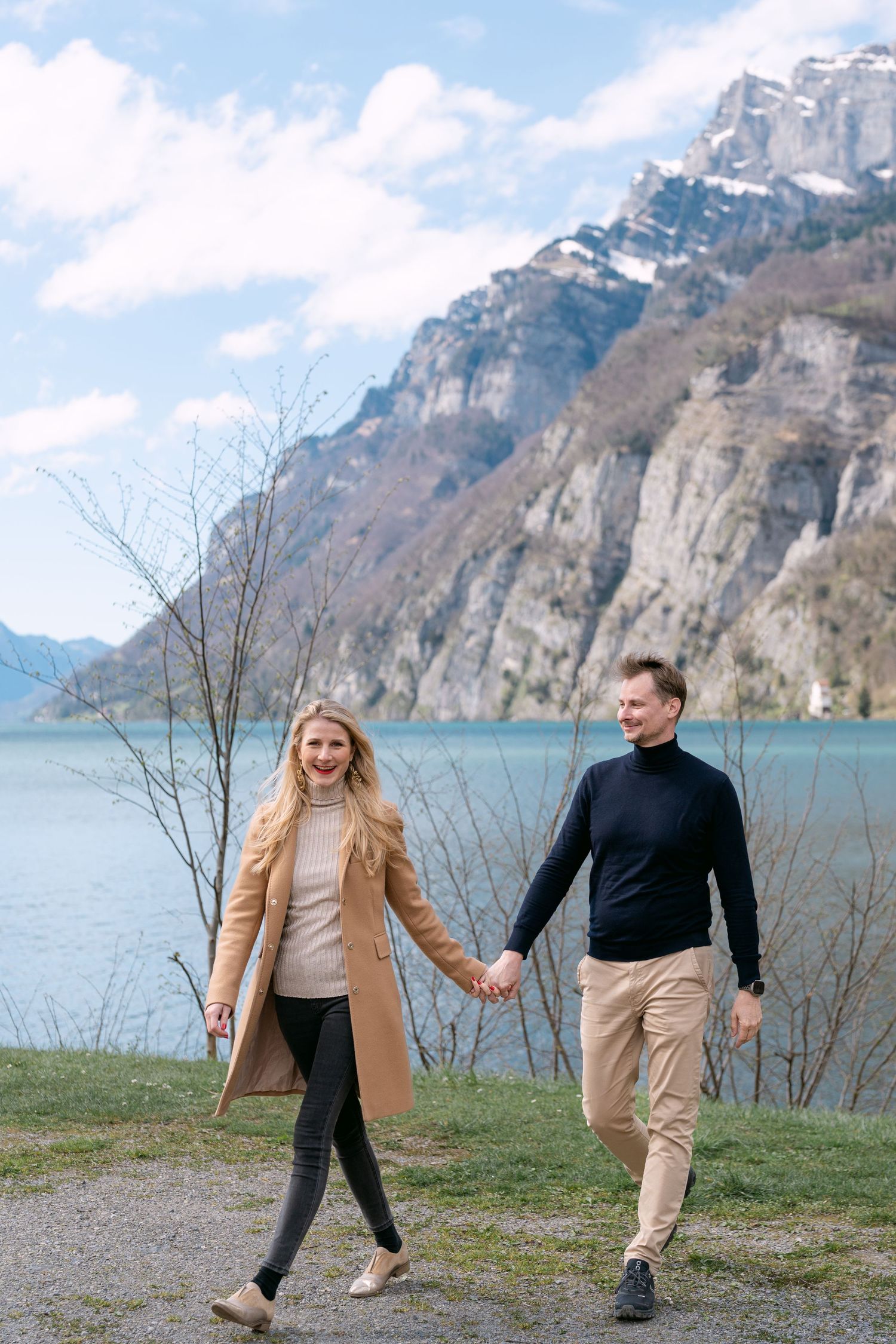 A couple walks hand in hand along lakeside path with dramatic mountain cliffs in background.