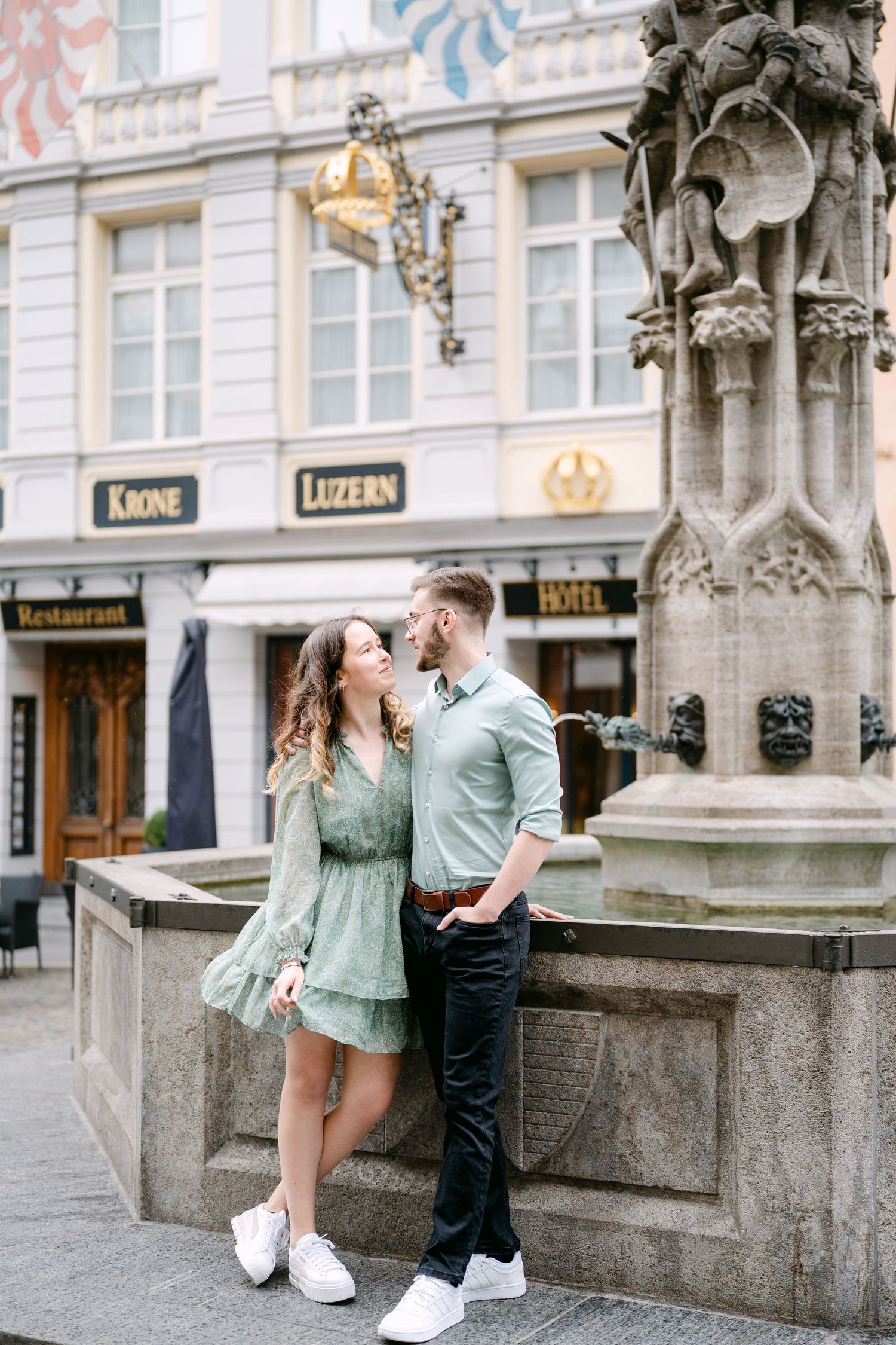 A couple poses romantically against ornate architecture in a European city square.