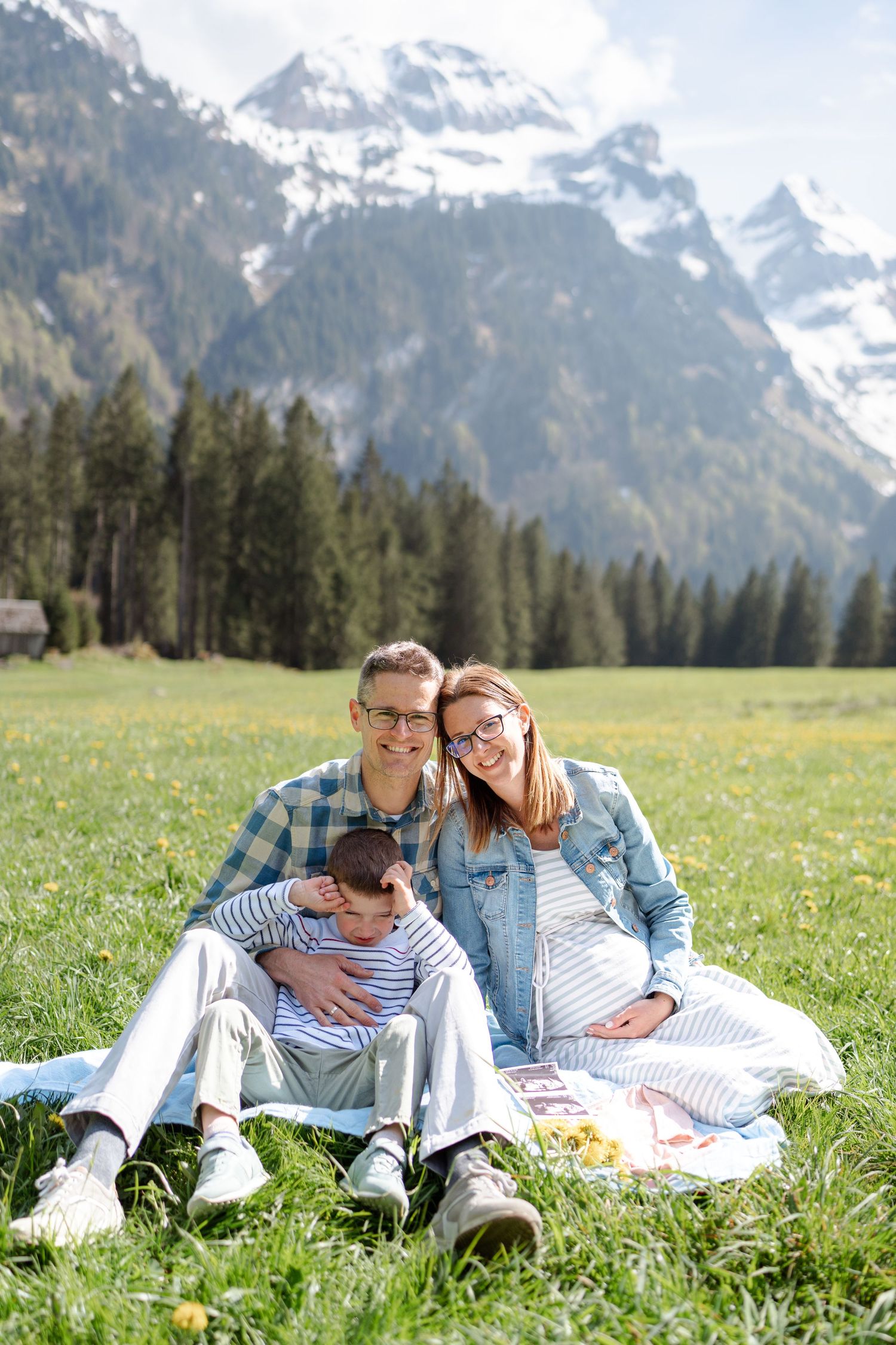 Family lounging together on white blanket in spring meadow with mountain vista.