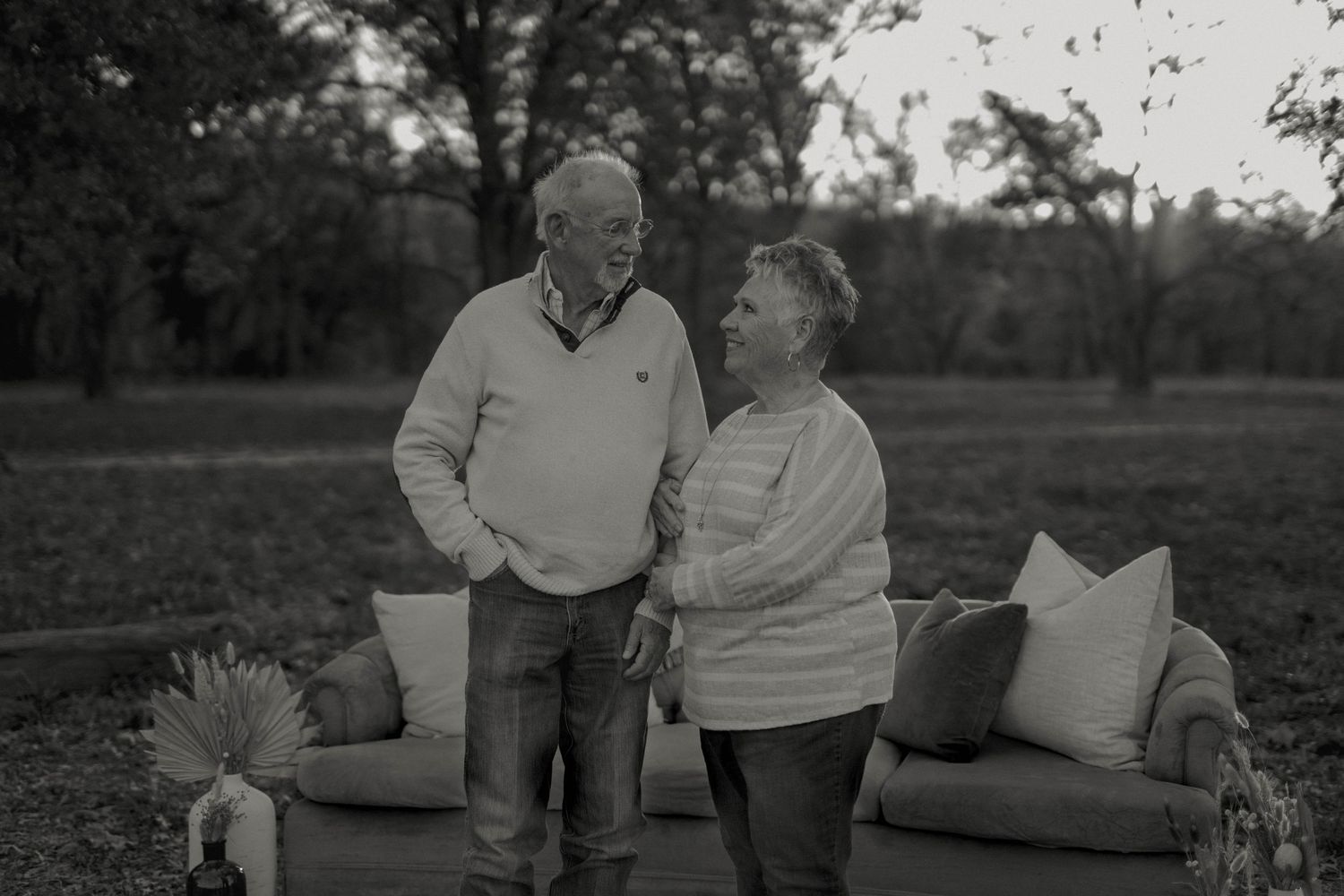 Elderly couple smiling and holding hands outdoors in a black and white photo surrounded by lush trees and patio furniture.