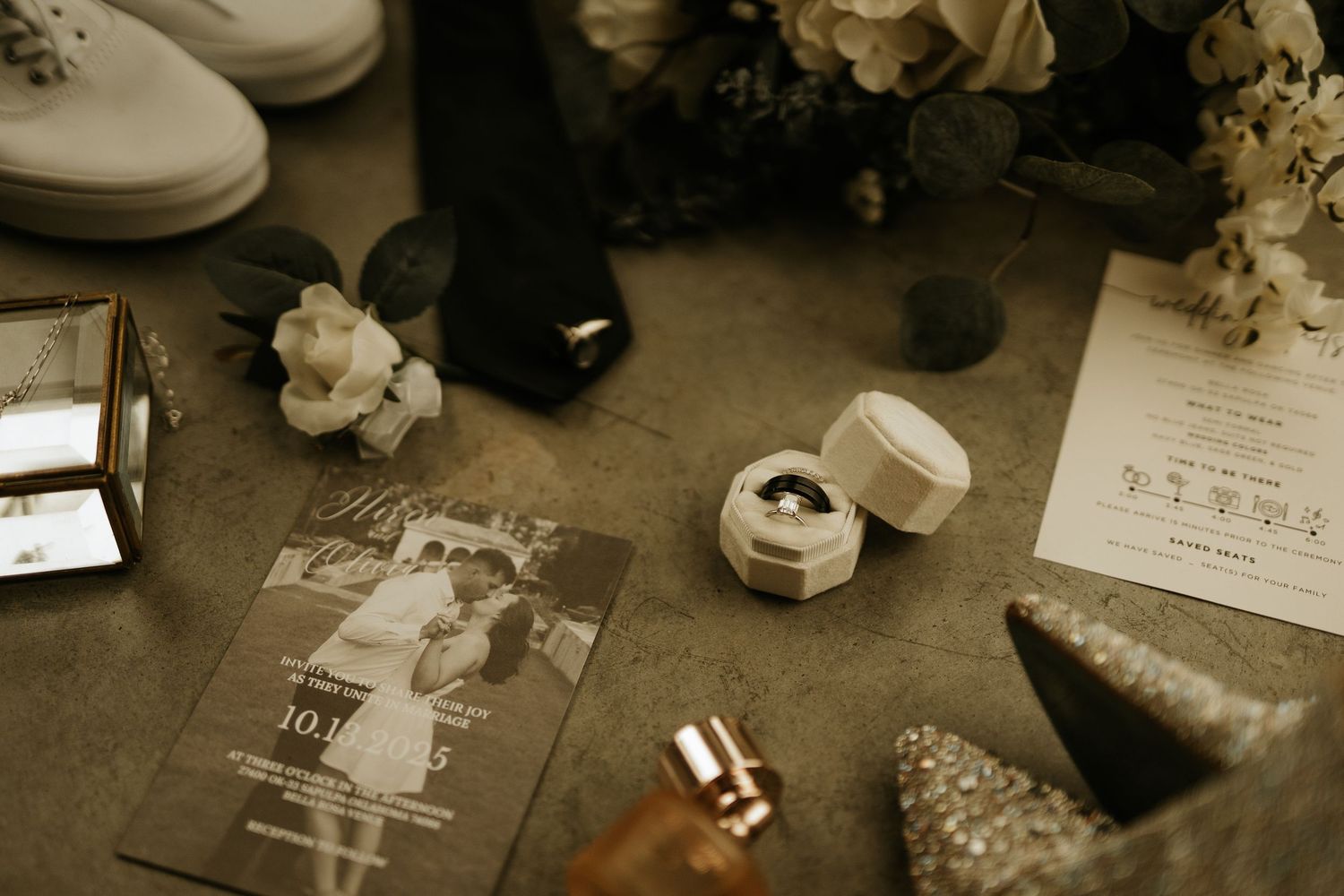 Wedding flat lay with ring box, invitation, perfume, bouquet, heels, and accessories on a gray surface.