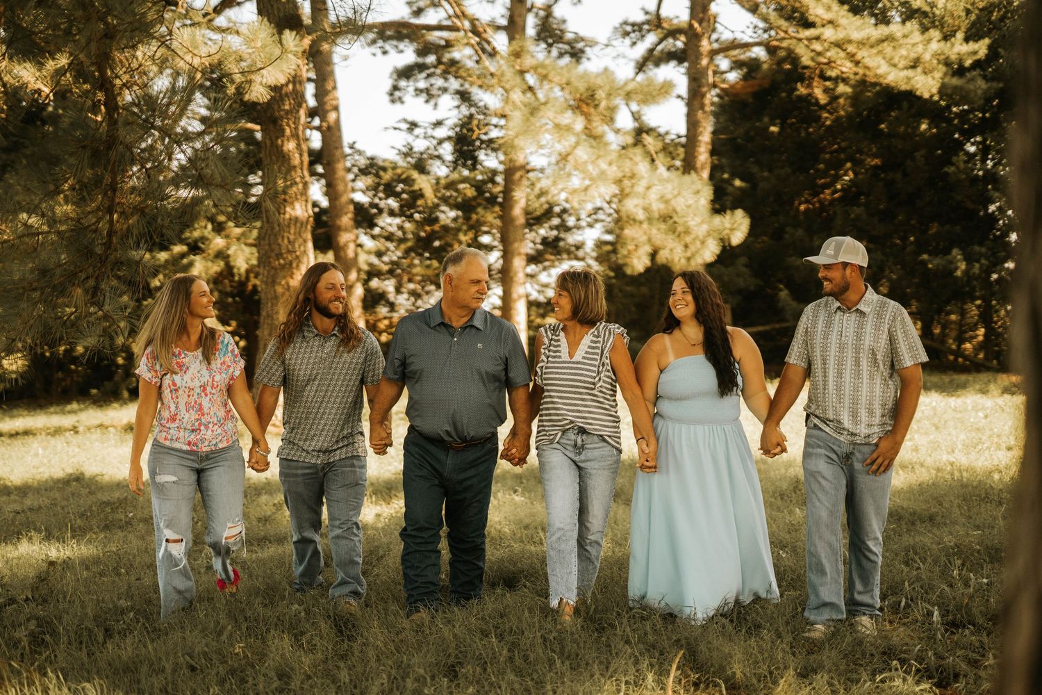 Six family members hold hands walking through a sunlit meadow surrounded by tall trees in a warm outdoor portrait.