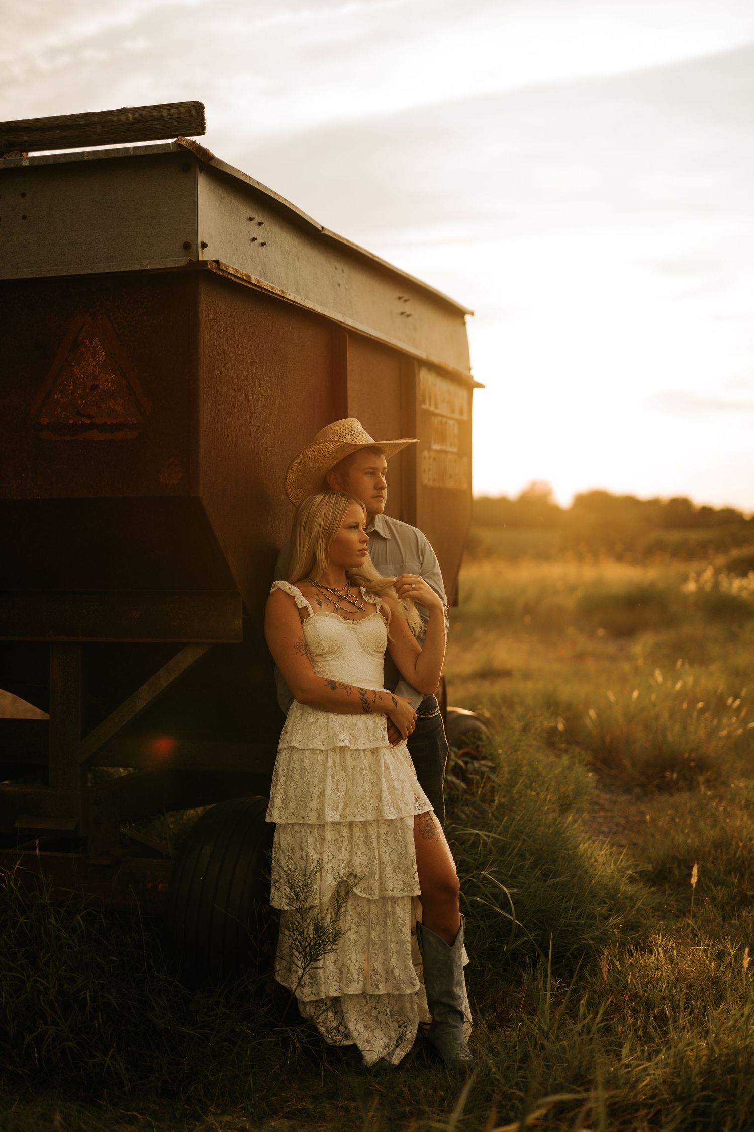 Couple in golden sunset light leaning against farm equipment, woman in white lace dress and boots, man in cowboy hat.