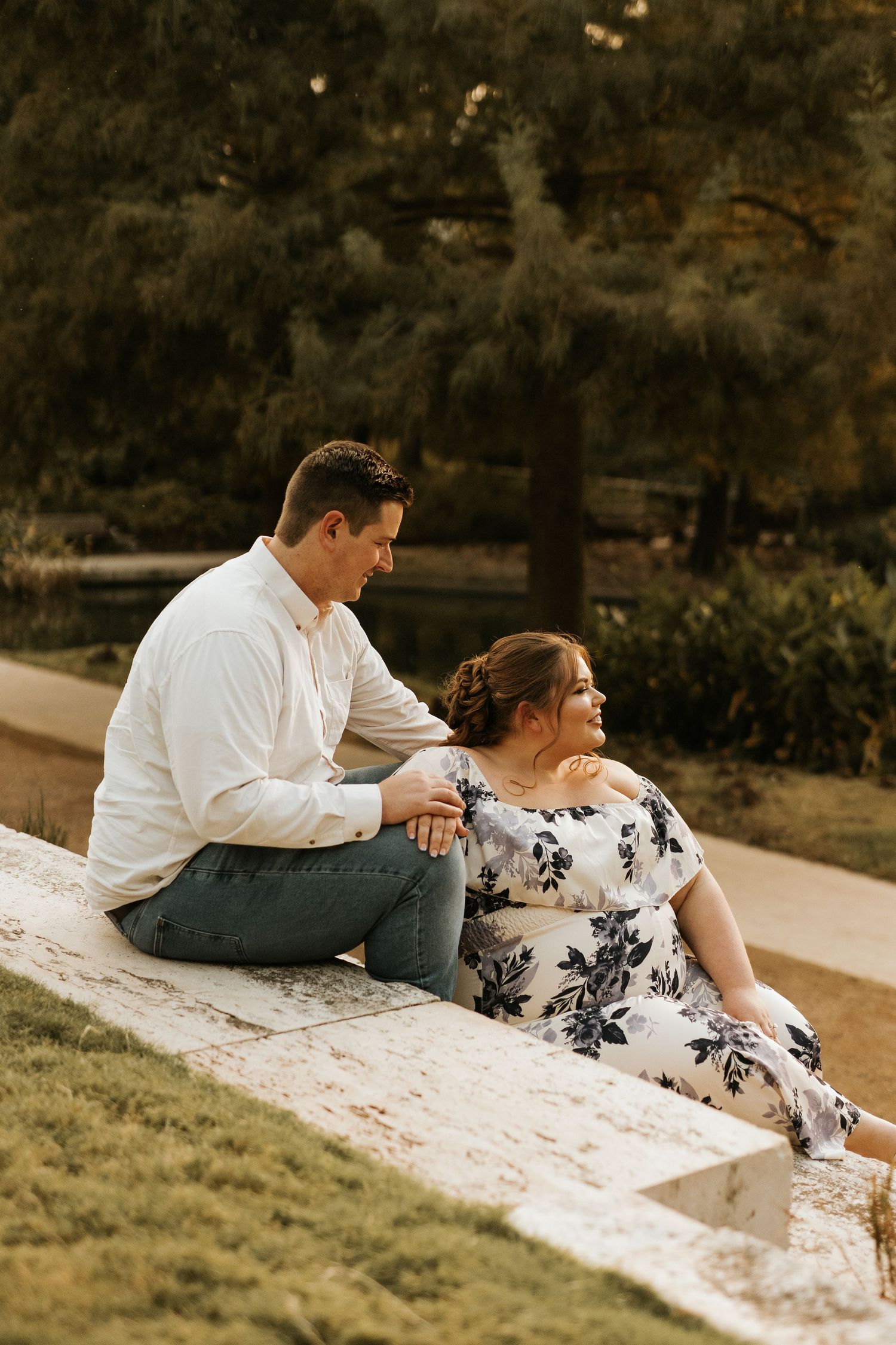 Couple sitting on stone steps in a park, man in white shirt and woman in floral dress, sharing a tender moment.