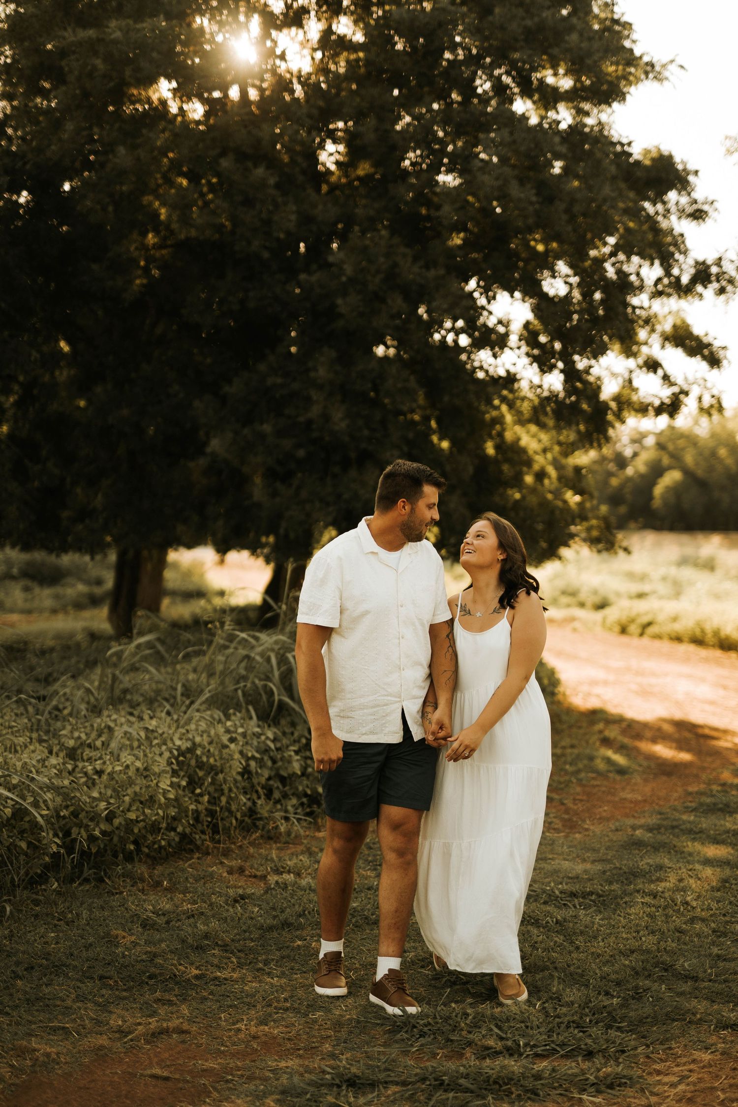 Couple holding hands and smiling at each other in a sunlit outdoor setting with lush green trees and a dirt path.