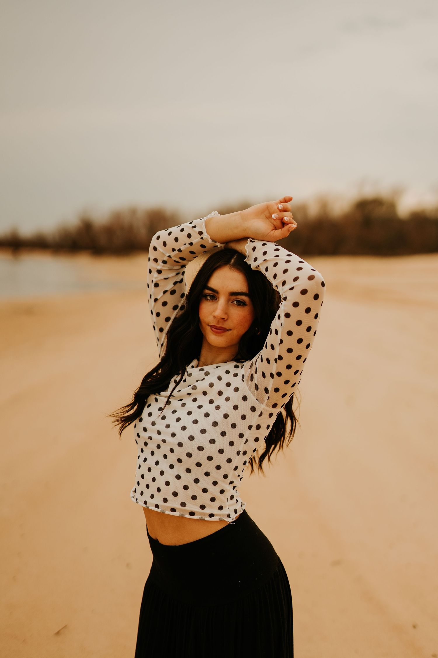 Young woman in polka dot crop top poses with arms raised on sandy beach under overcast sky.