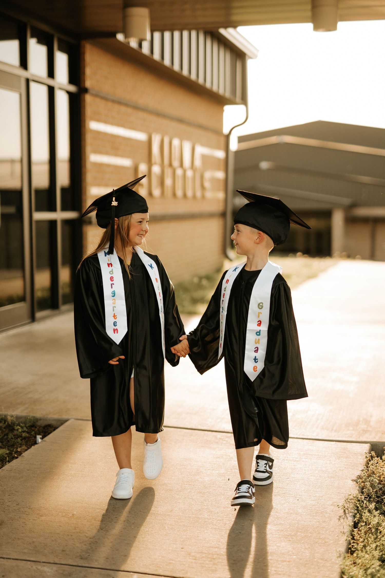 Two graduates in matching caps and gowns walk together on school sidewalk.