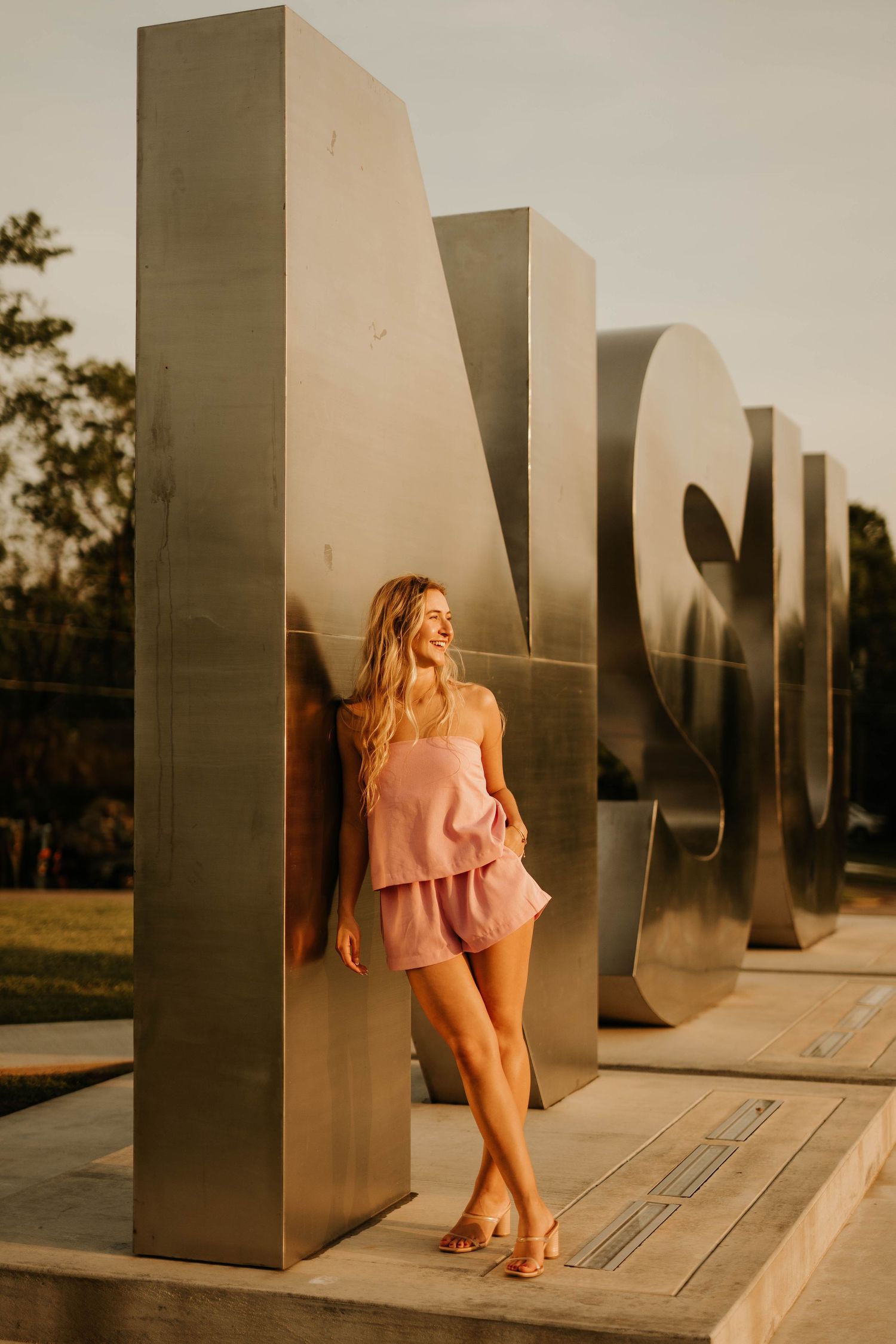 Someone in a pink outfit poses against modern metallic sculptural columns at sunset.