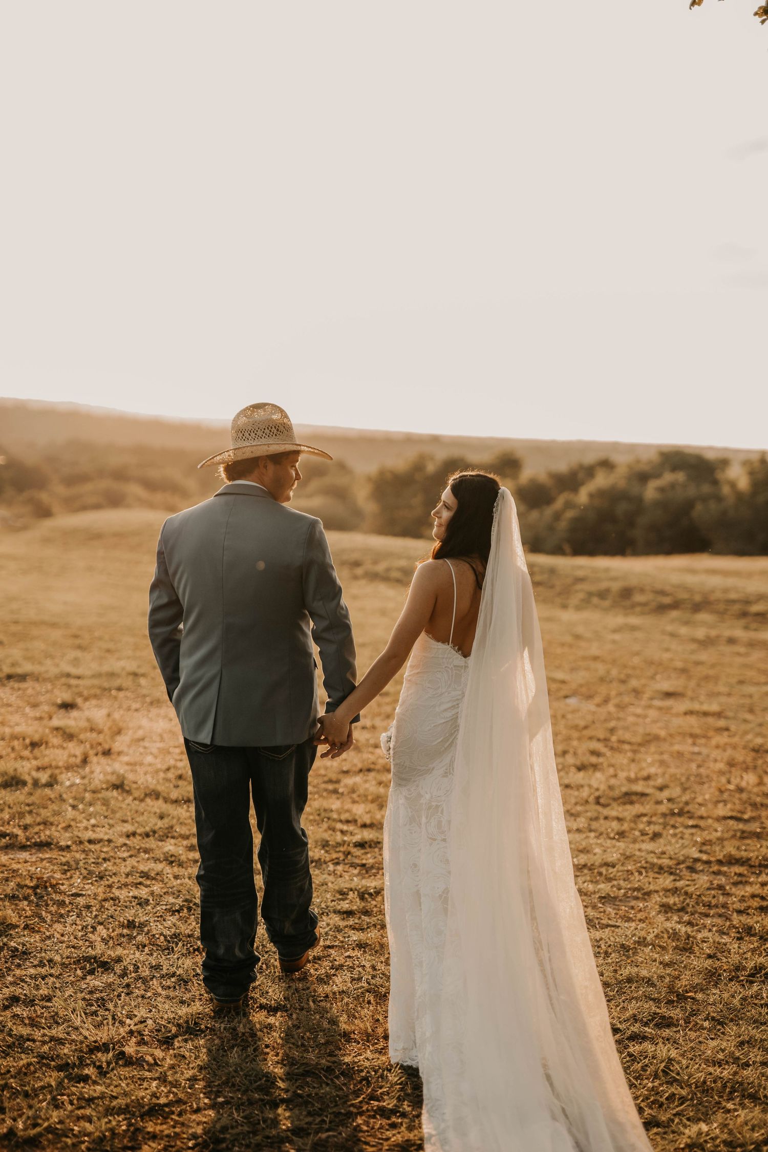 A couple holding hands at sunset in a field with the bride wearing a flowing white wedding gown.