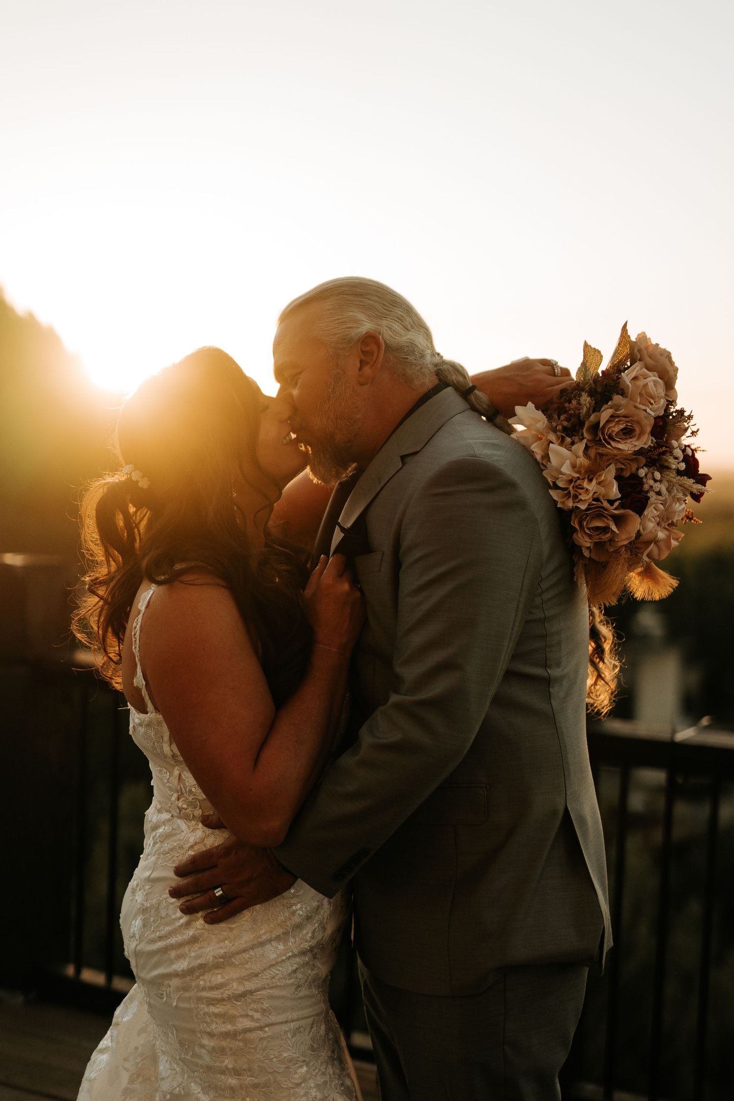 A bride and groom share a romantic kiss at golden hour sunset, with the bride holding a floral bouquet.