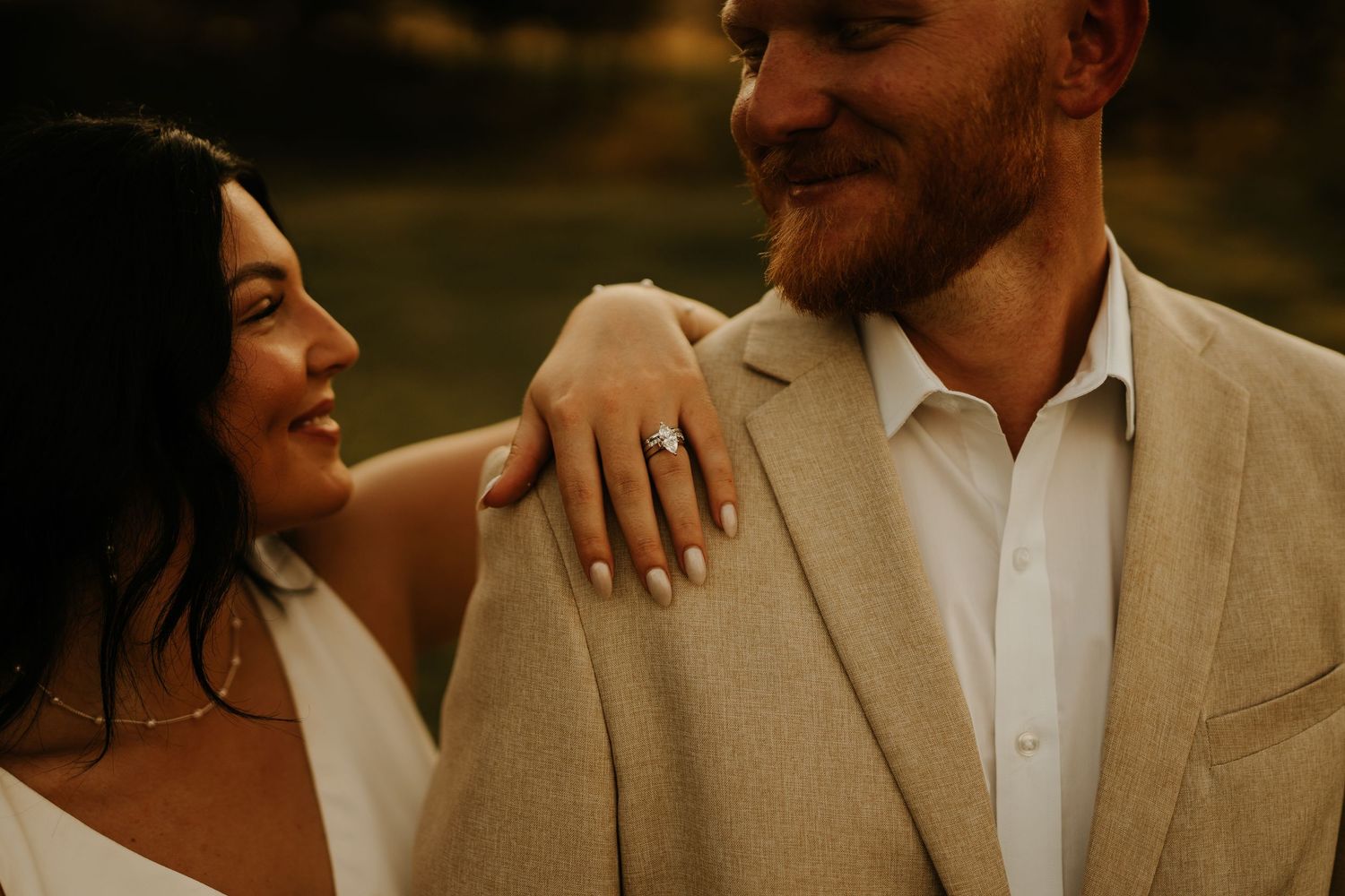 Smiling bride shows off engagement ring while leaning on groom in tan suit during romantic outdoor photo session.
