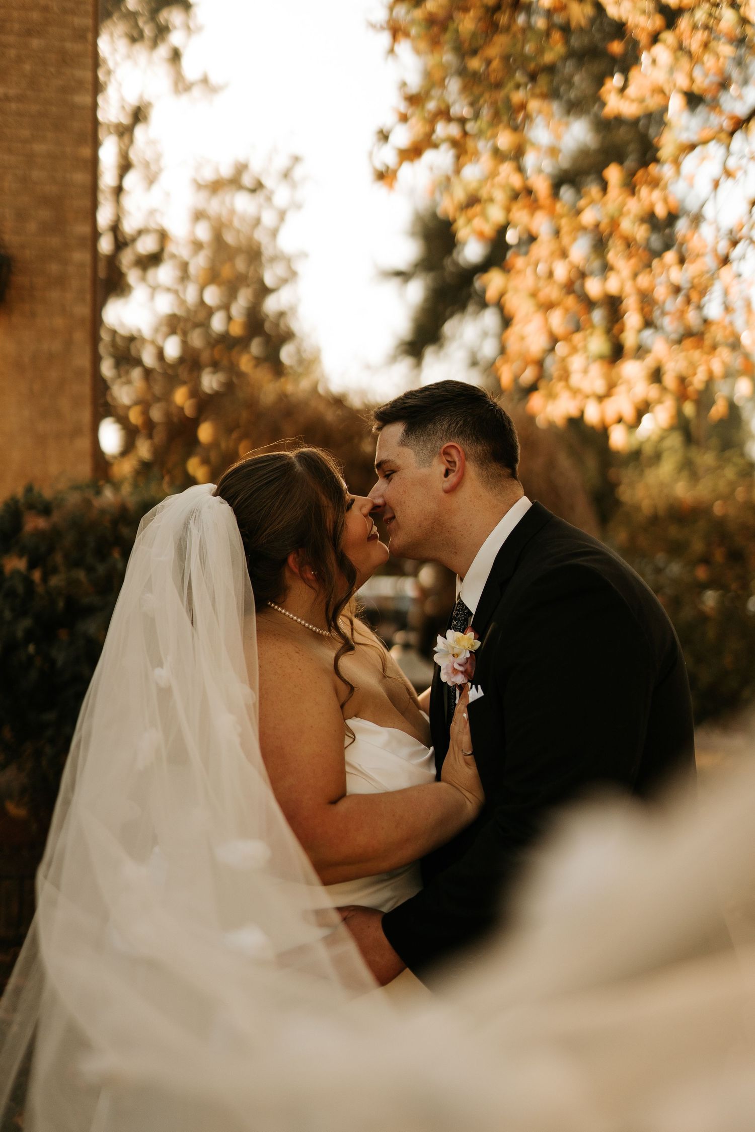 A bride and groom share a romantic almost-kiss outdoors, surrounded by golden autumn foliage in warm evening light.