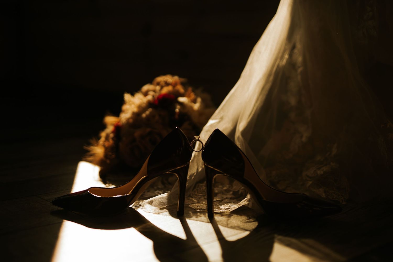 Elegant bridal heels and floral hair accessory resting on a dark floor in dramatic golden light beside a white wedding dress.