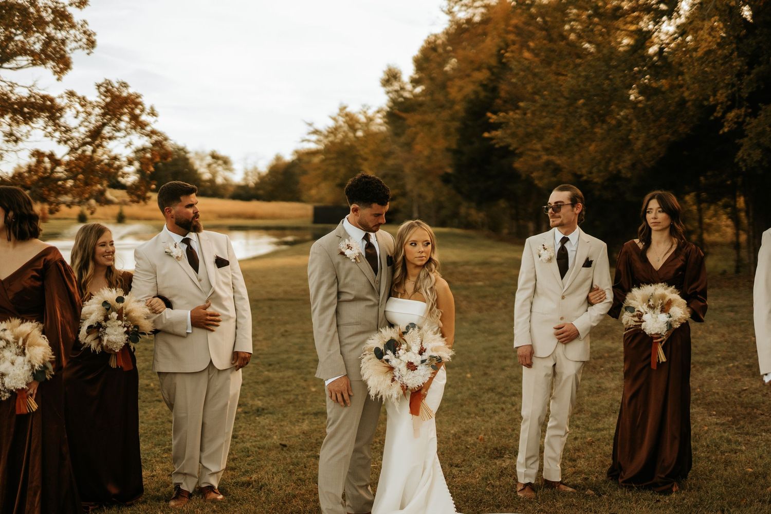 Wedding party poses outdoors in autumn, bride holding bouquet, groom in tan suit, groomsmen and bridesmaids in brown attire.
