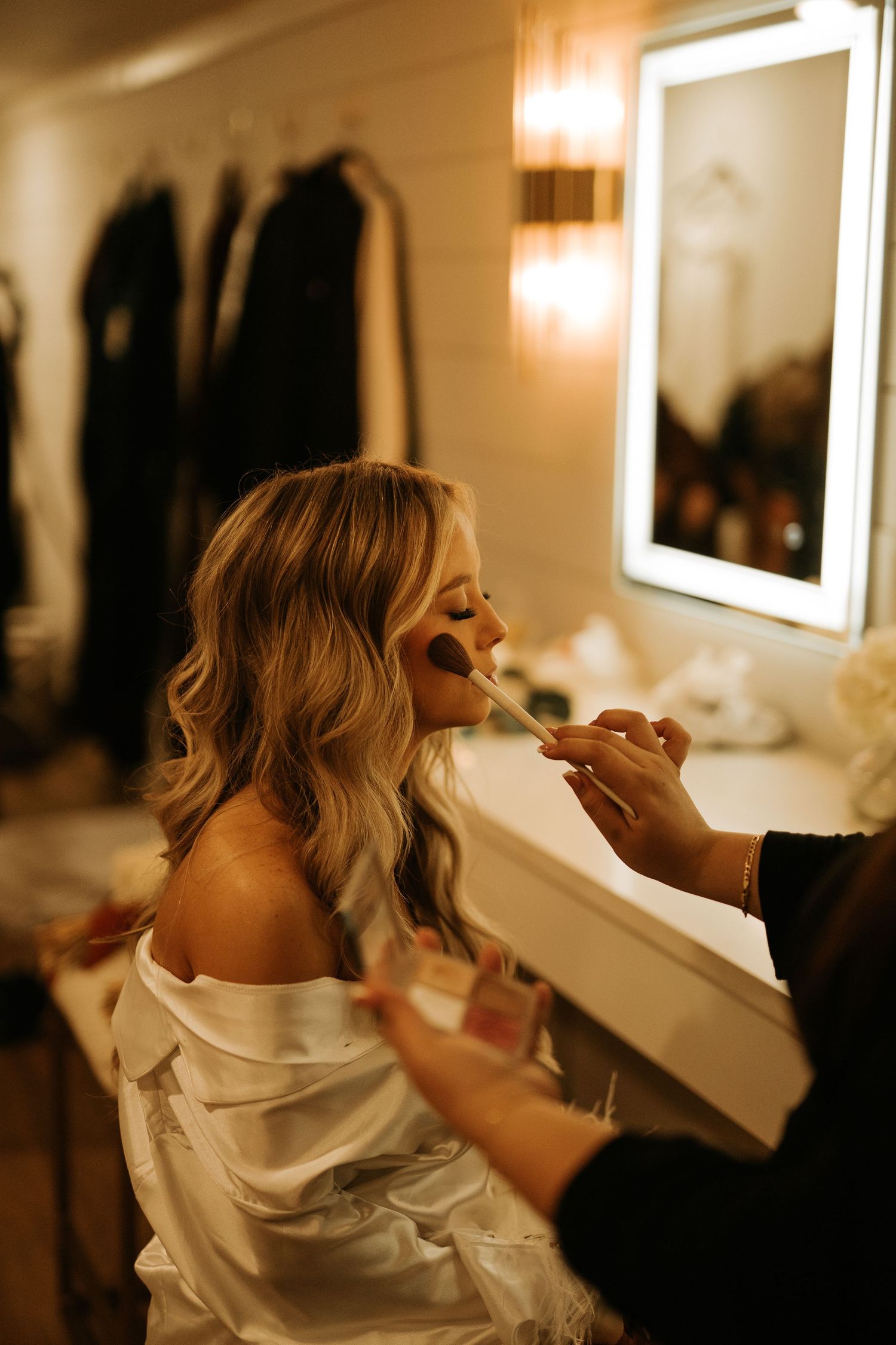 Blonde bride in white robe getting makeup applied by artist near illuminated vanity mirror.
