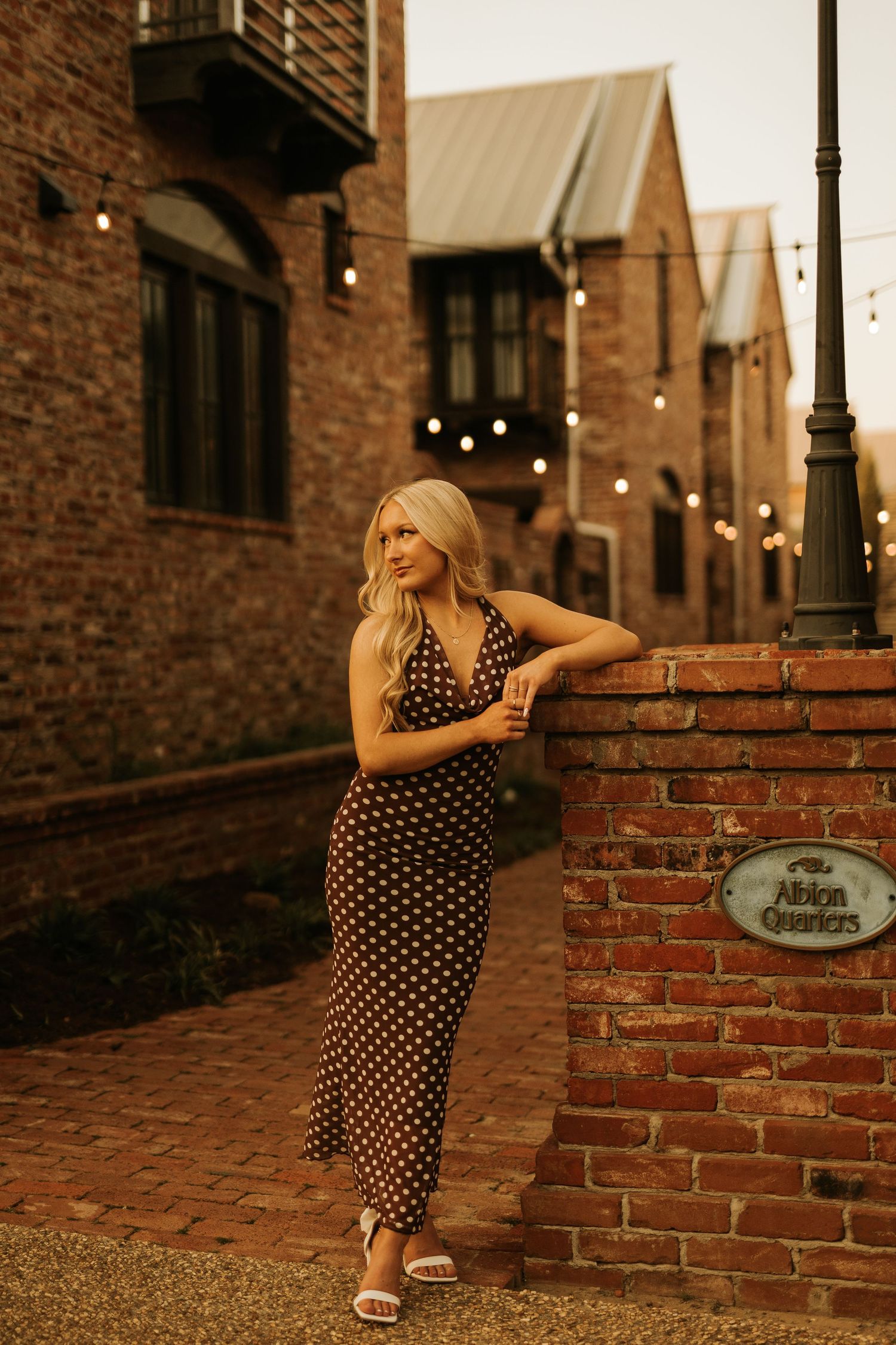 Blonde woman in polka dot maxi dress leaning on brick wall at Albion Quarters during golden hour.