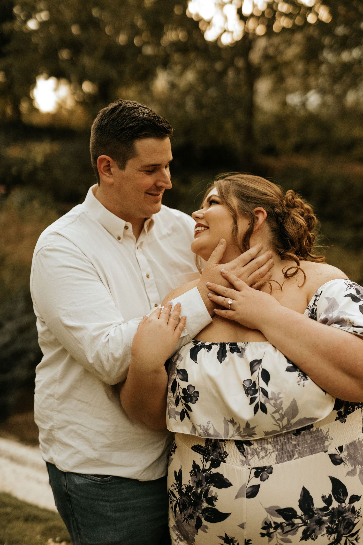 A couple shares a romantic moment outdoors, man in white shirt tenderly holds woman's face as she smiles in floral dress.