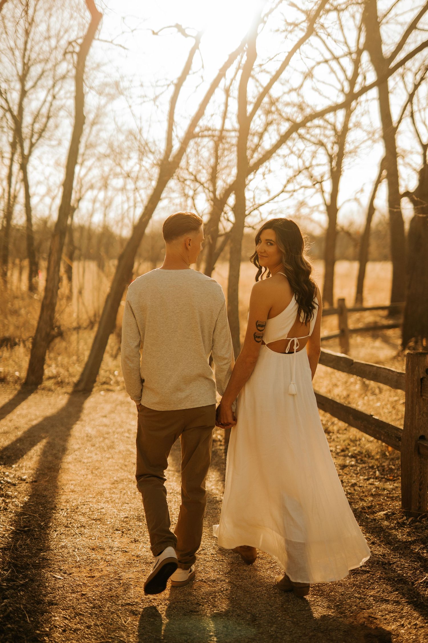 Couple holding hands walking on a sunlit path by a wooden fence, woman in white dress glancing back at golden hour.
